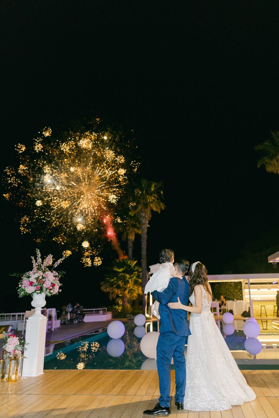 Wedding couple watching fireworks display from outdoor reception venue at night.