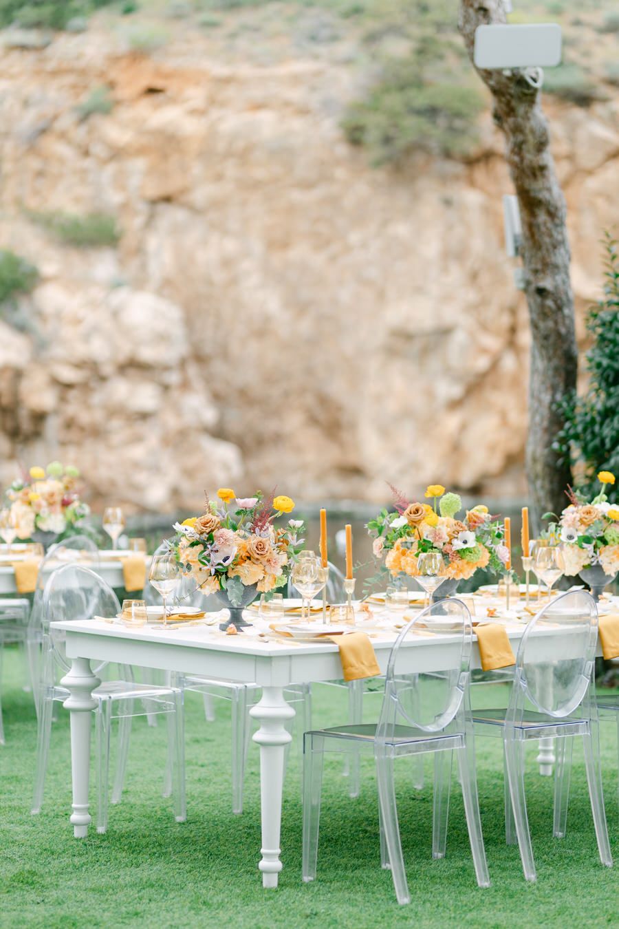 Outdoor wedding setup with white dining tables and ghost chairs against rocky landscape.