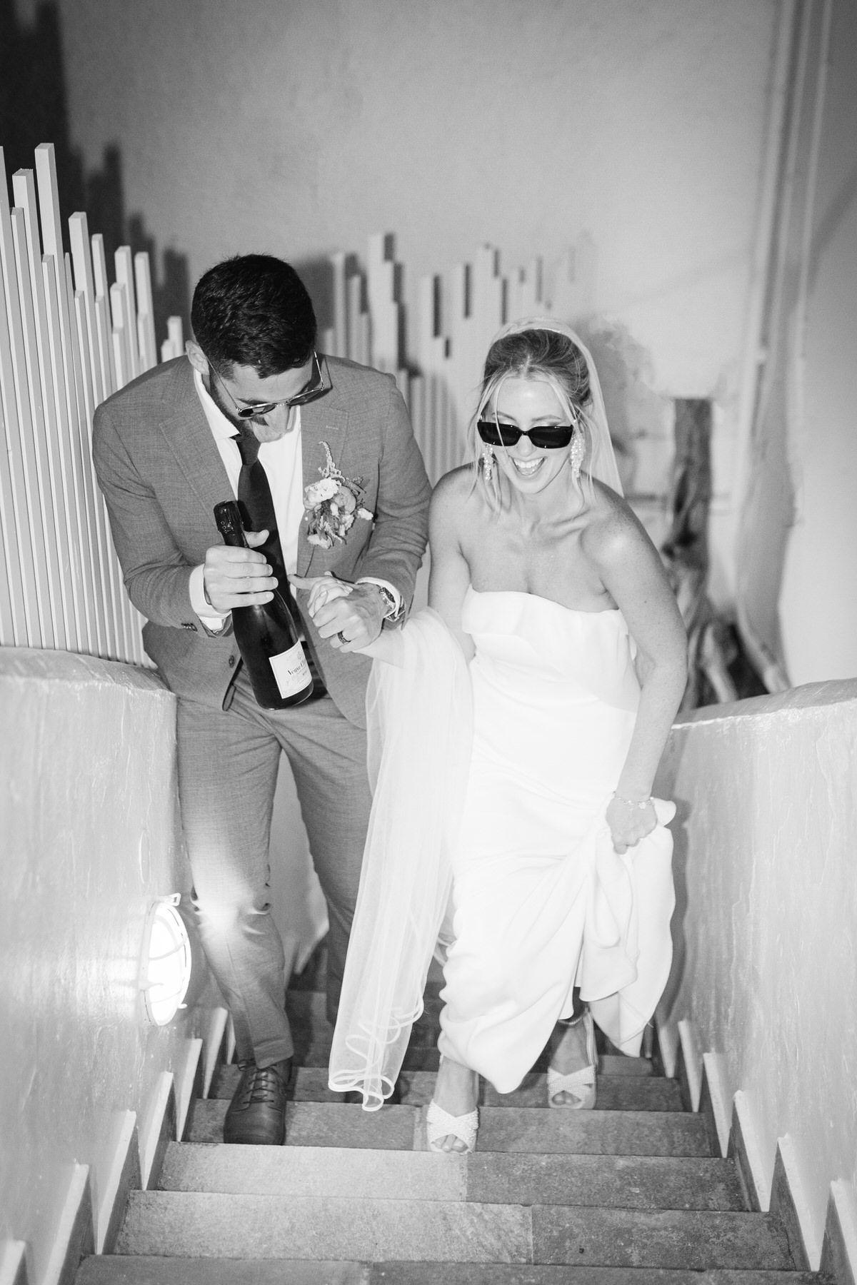 A couple in formal attire descends stairs with champagne, bride wearing sunglasses and white dress, groom in gray suit.