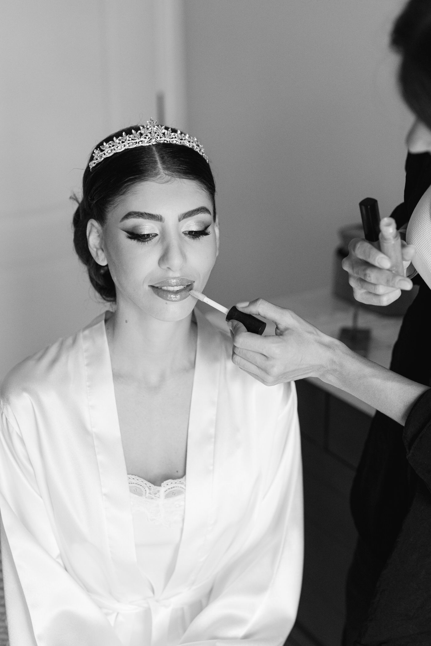 Black and white photo showing a bride getting her makeup done while wearing a white silk robe and delicate tiara.