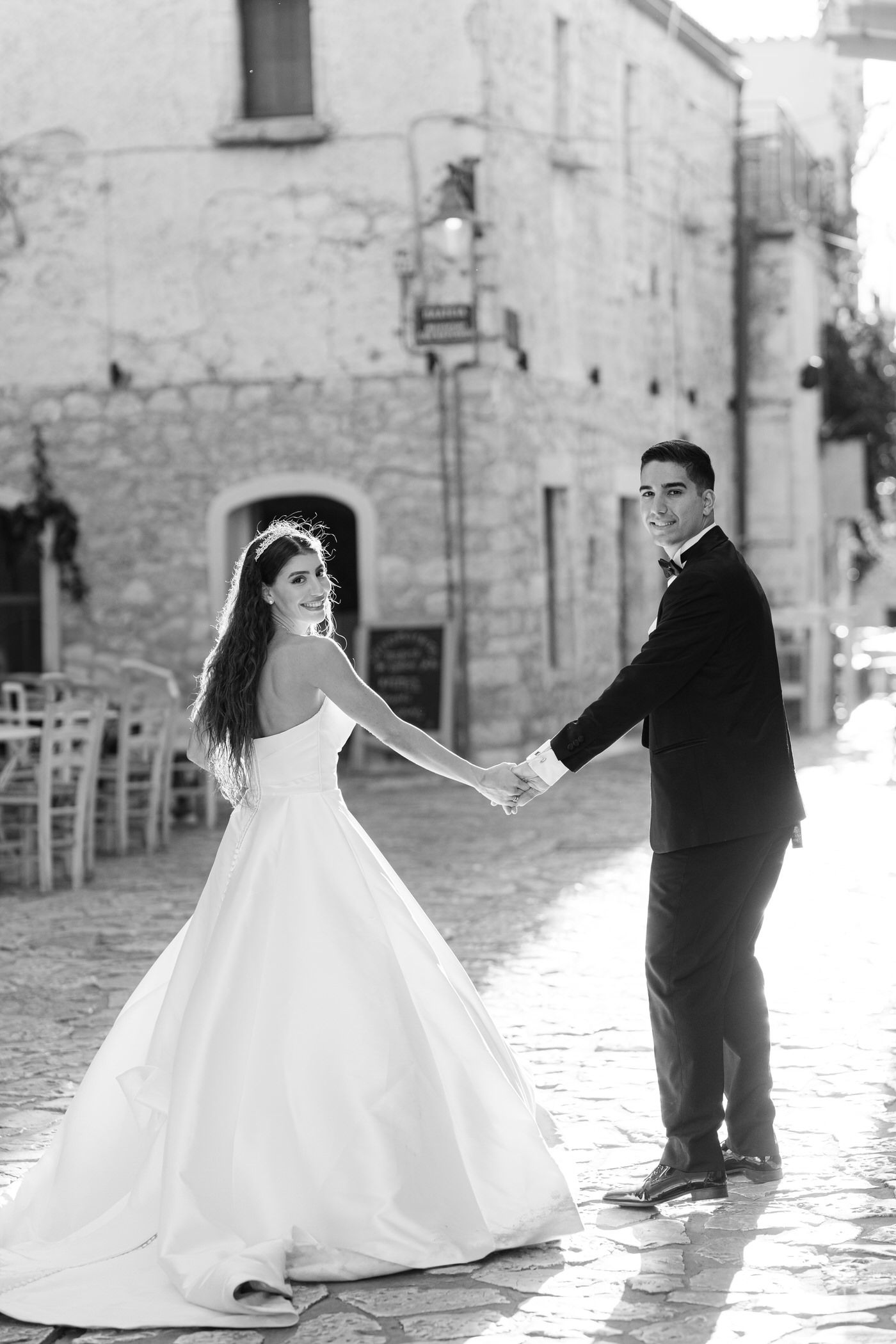 A romantic black and white wedding photo of a couple in formal attire holding hands on a cobblestone street in Italy.