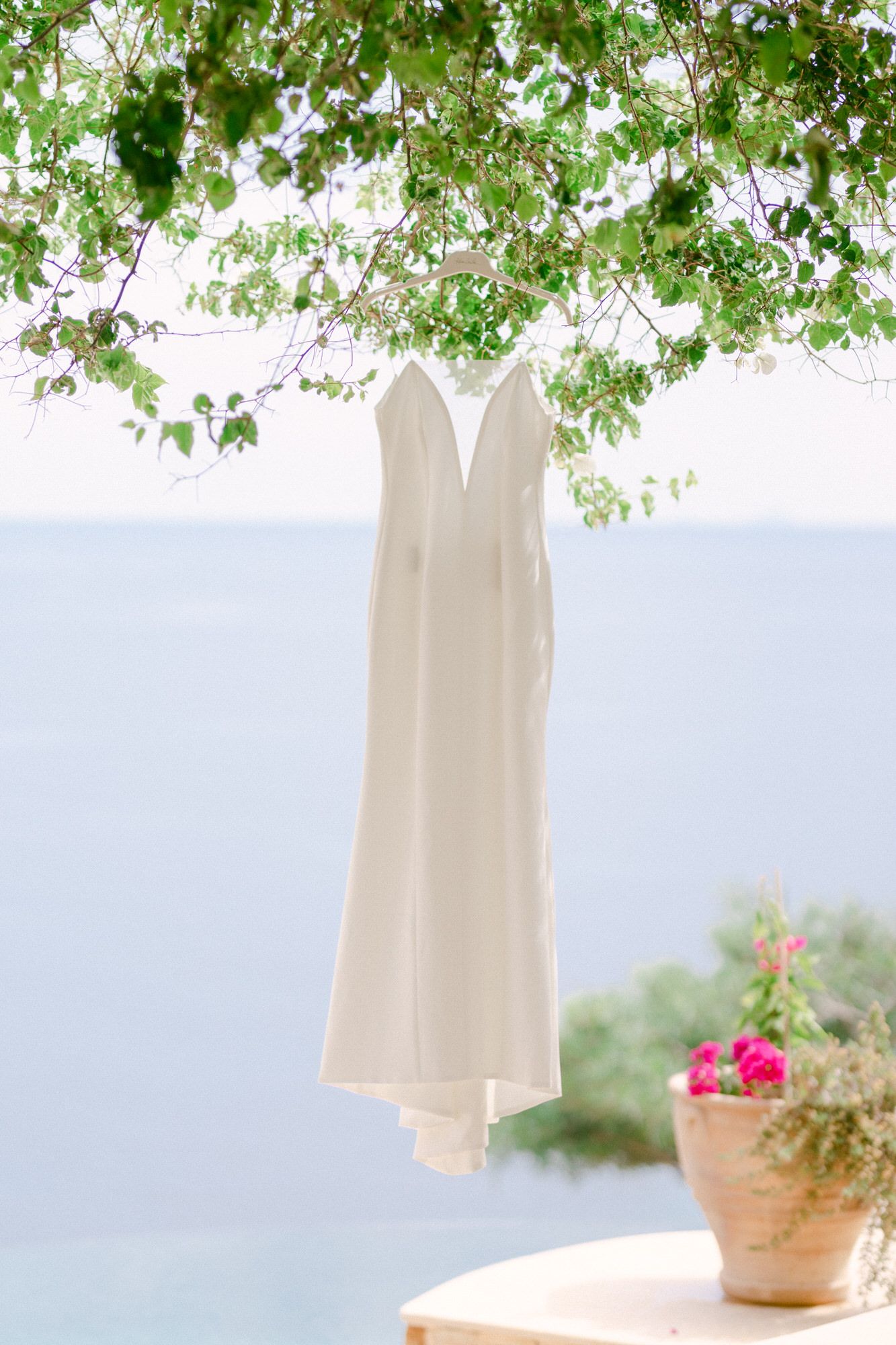 An elegant white wedding dress hanging from tree branches with ocean view in background.