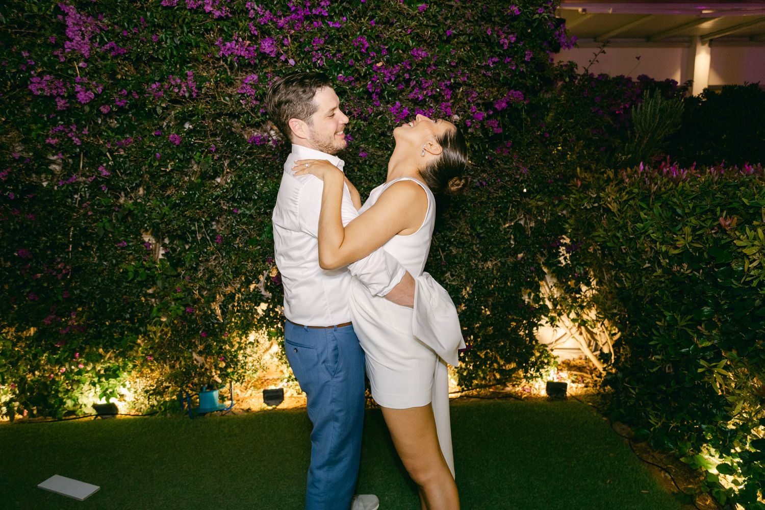 A dancing couple in white attire share an intimate moment in a garden illuminated by warm ground lights at night.