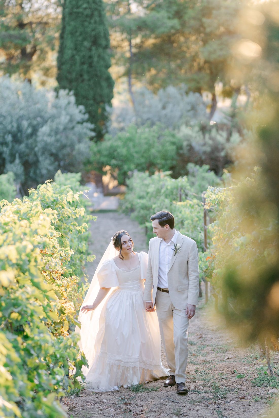 A couple in wedding attire walk together through a sun-drenched vineyard path with lush greenery and cypress trees.