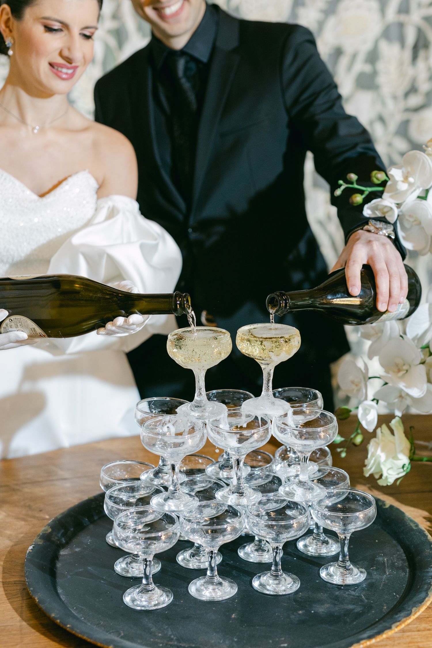 Champagne being poured into vintage coupe glasses arranged in a tower on a black tray at an elegant wedding celebration.