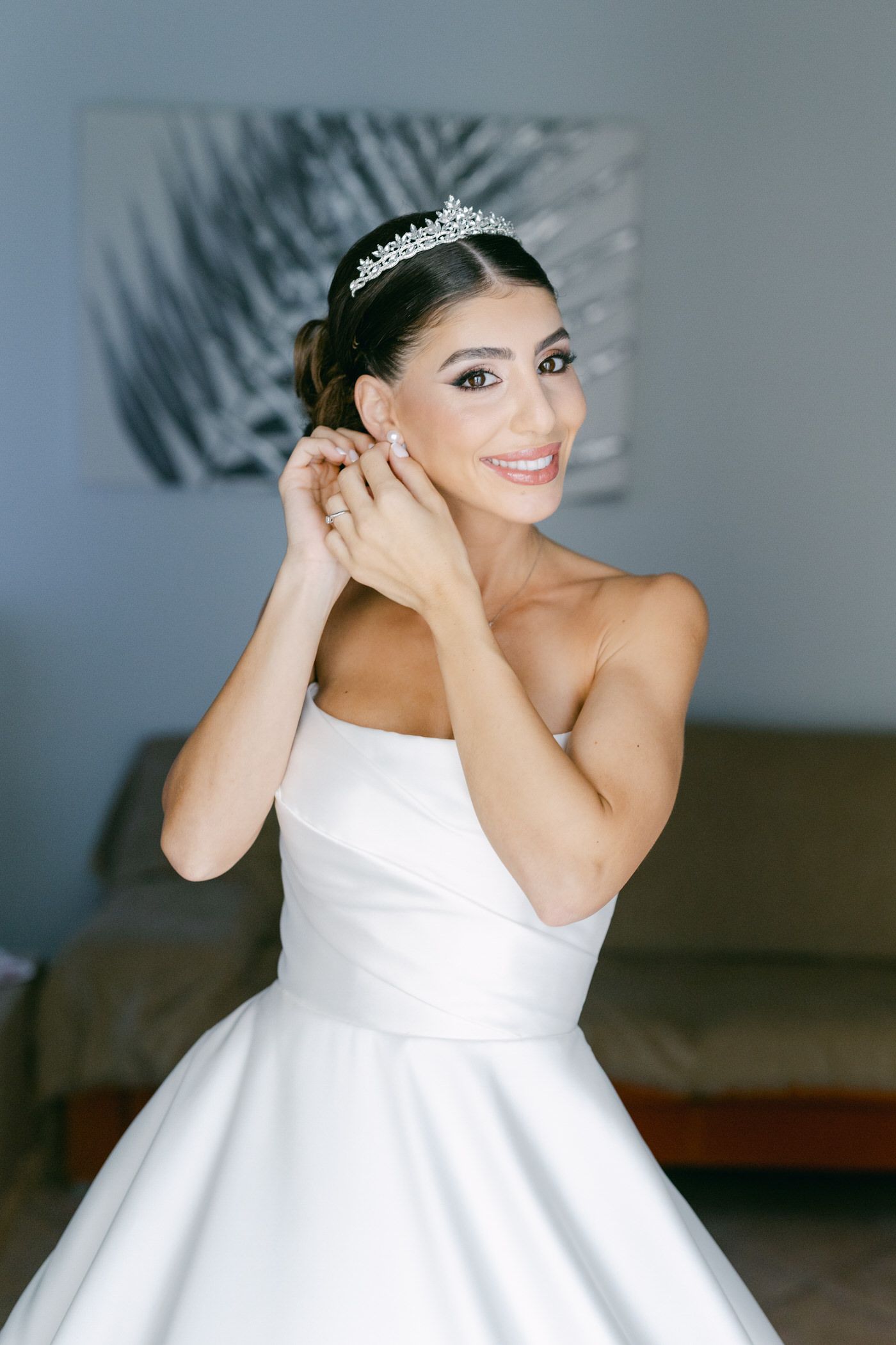 Bride wearing elegant white strapless ballgown and crystal crown smiles while putting on earrings.