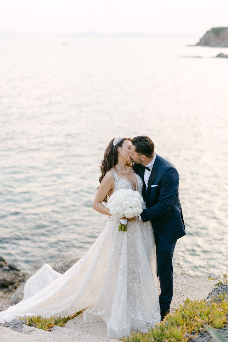 Romantic coastal wedding photo of couple embracing at sunset with bride in flowing lace gown holding white bouquet.