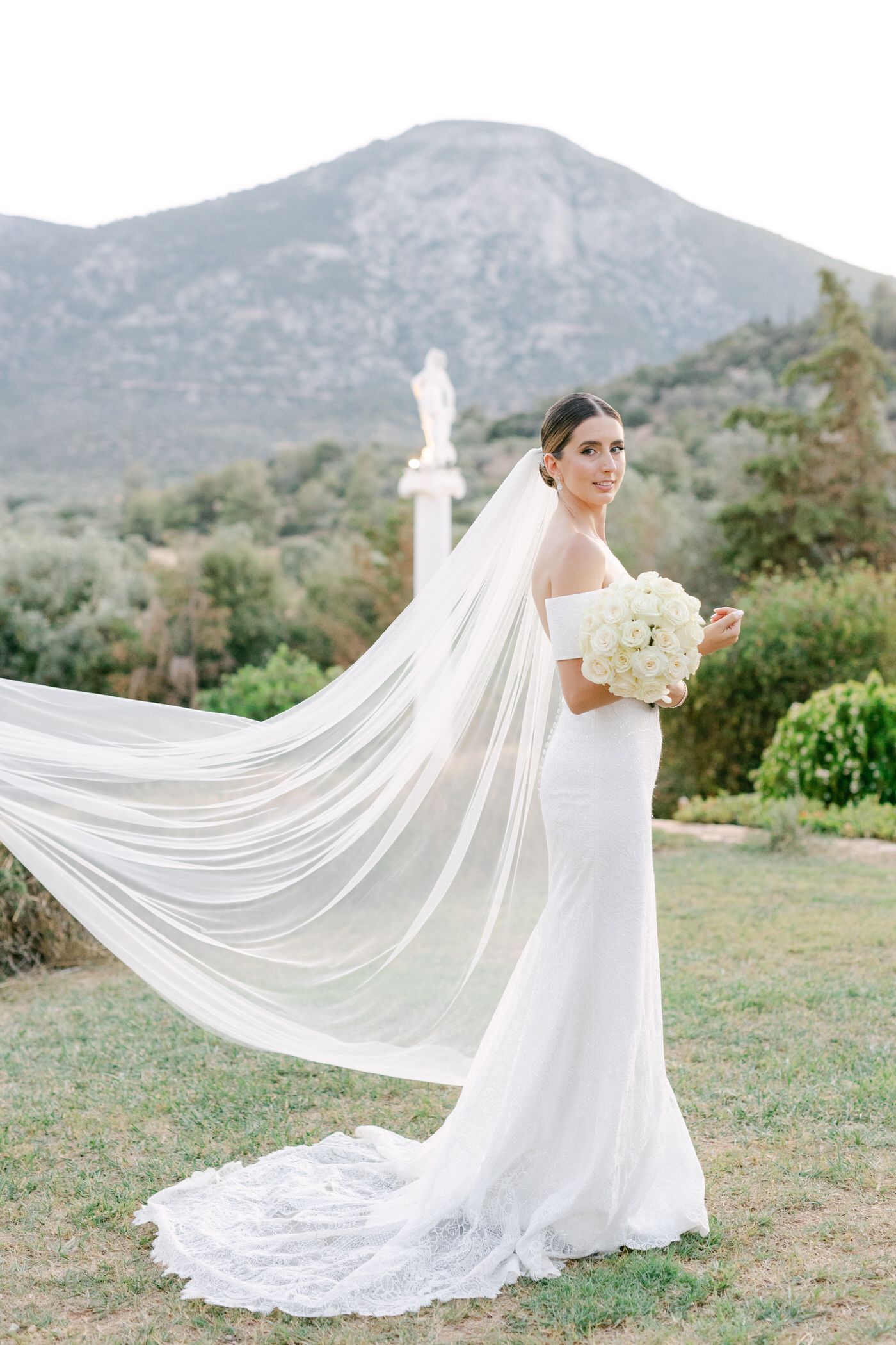 A bride in a form-fitting white gown with flowing cathedral veil poses outdoors with mountains in the background.