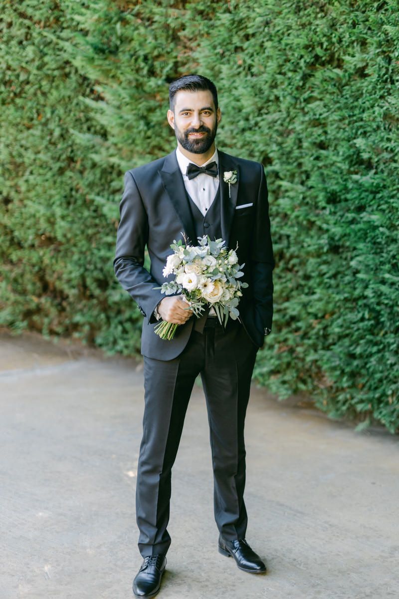 A well-dressed person in a black tuxedo holds a delicate bouquet of flowers while standing on a pathway with green hedges.