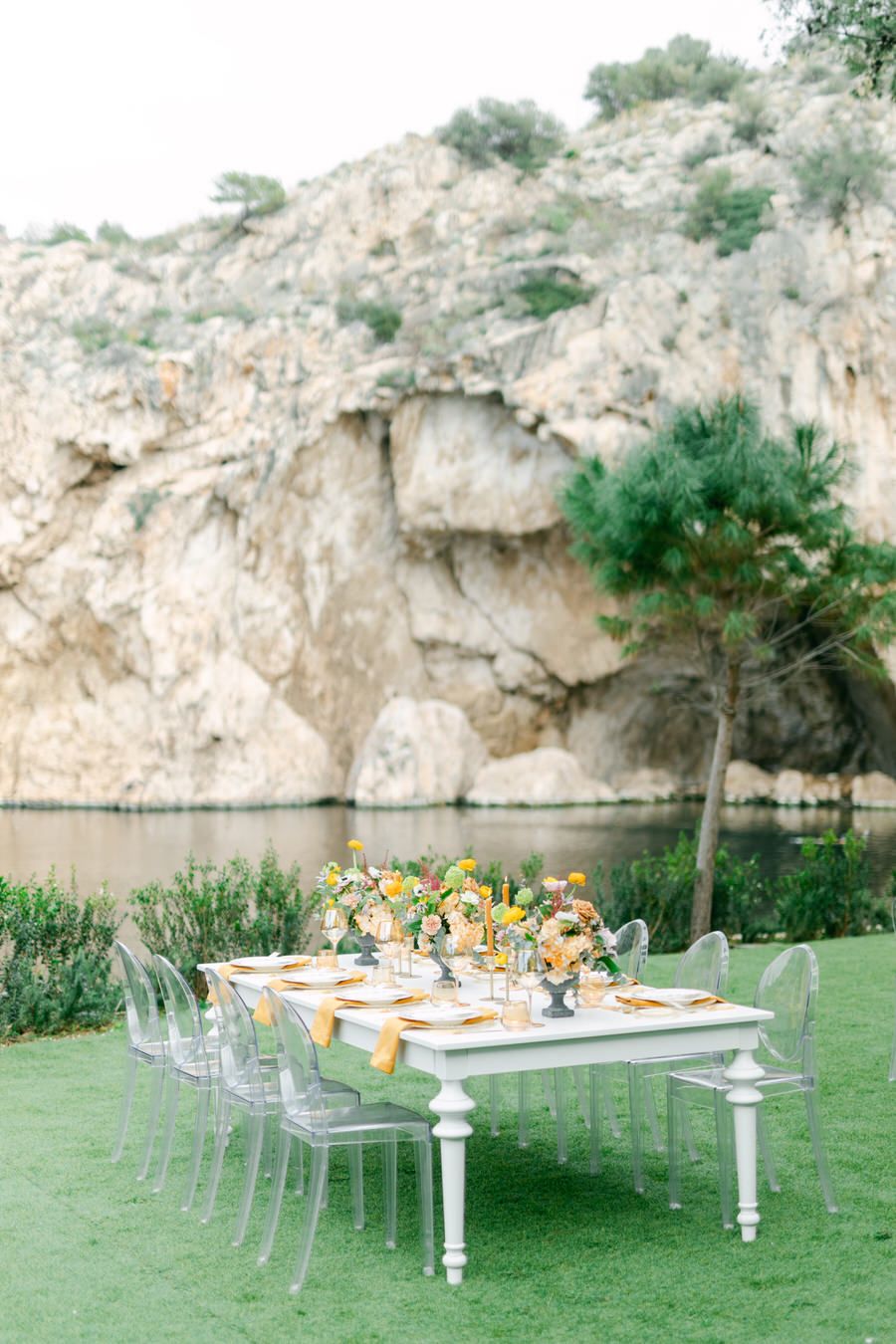 Elegant outdoor dining table setup with ghost chairs against dramatic rocky cliff landscape and water backdrop.