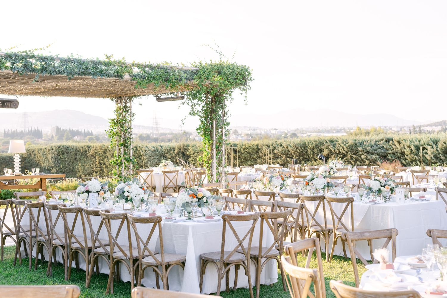 Elegant outdoor wedding reception setup with wooden cross-back chairs and white tables under a greenery-covered pergola.