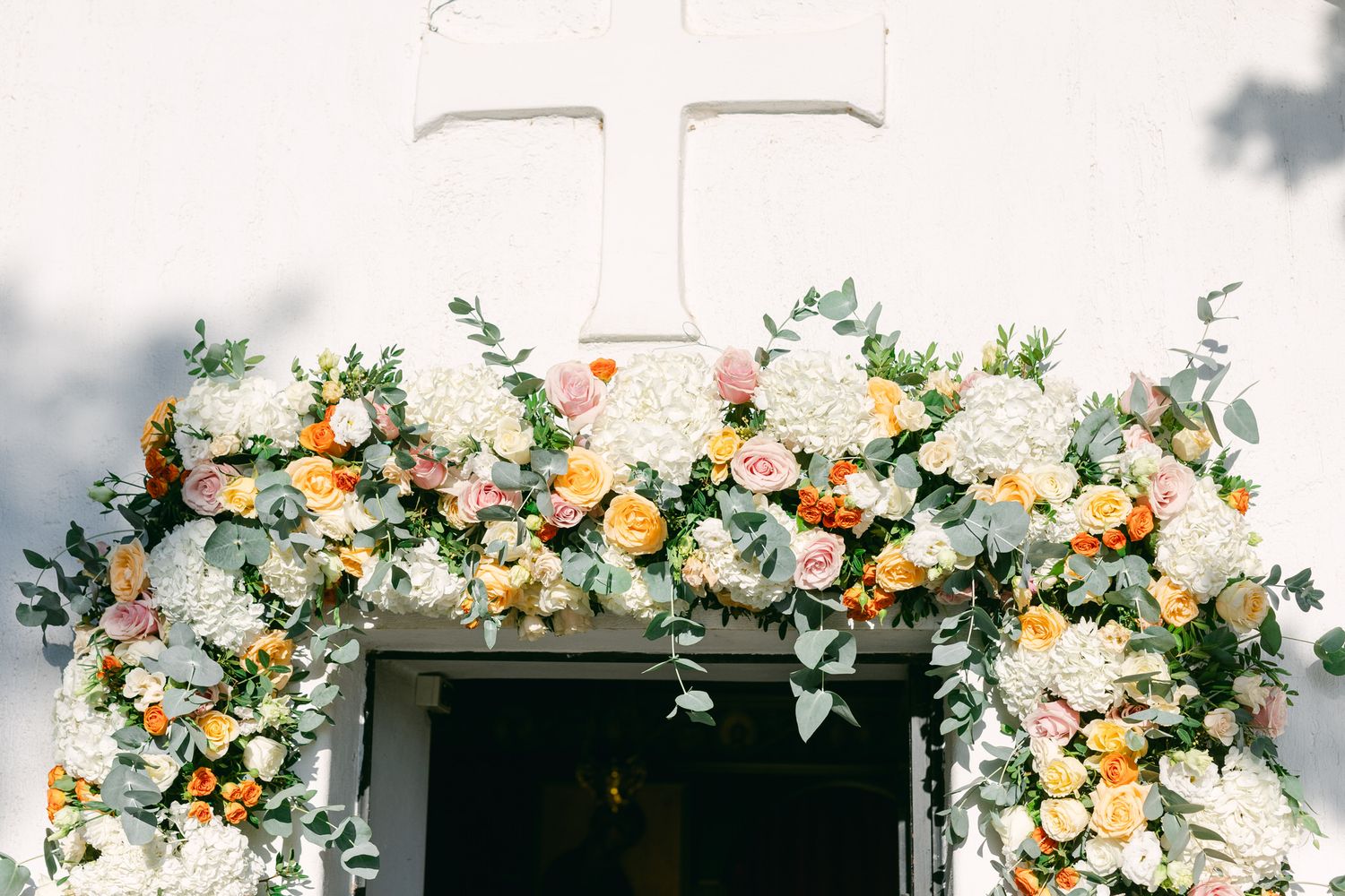 Floral wedding arch with white roses, peach ranunculus, and greenery frames a church doorway in sunlight.