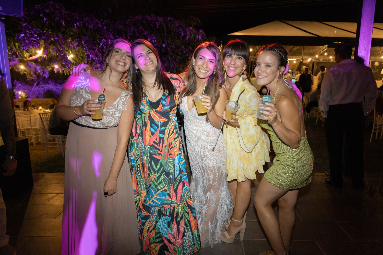 Party guests in colorful dresses pose together with drinks at an evening reception with purple lighting effects.