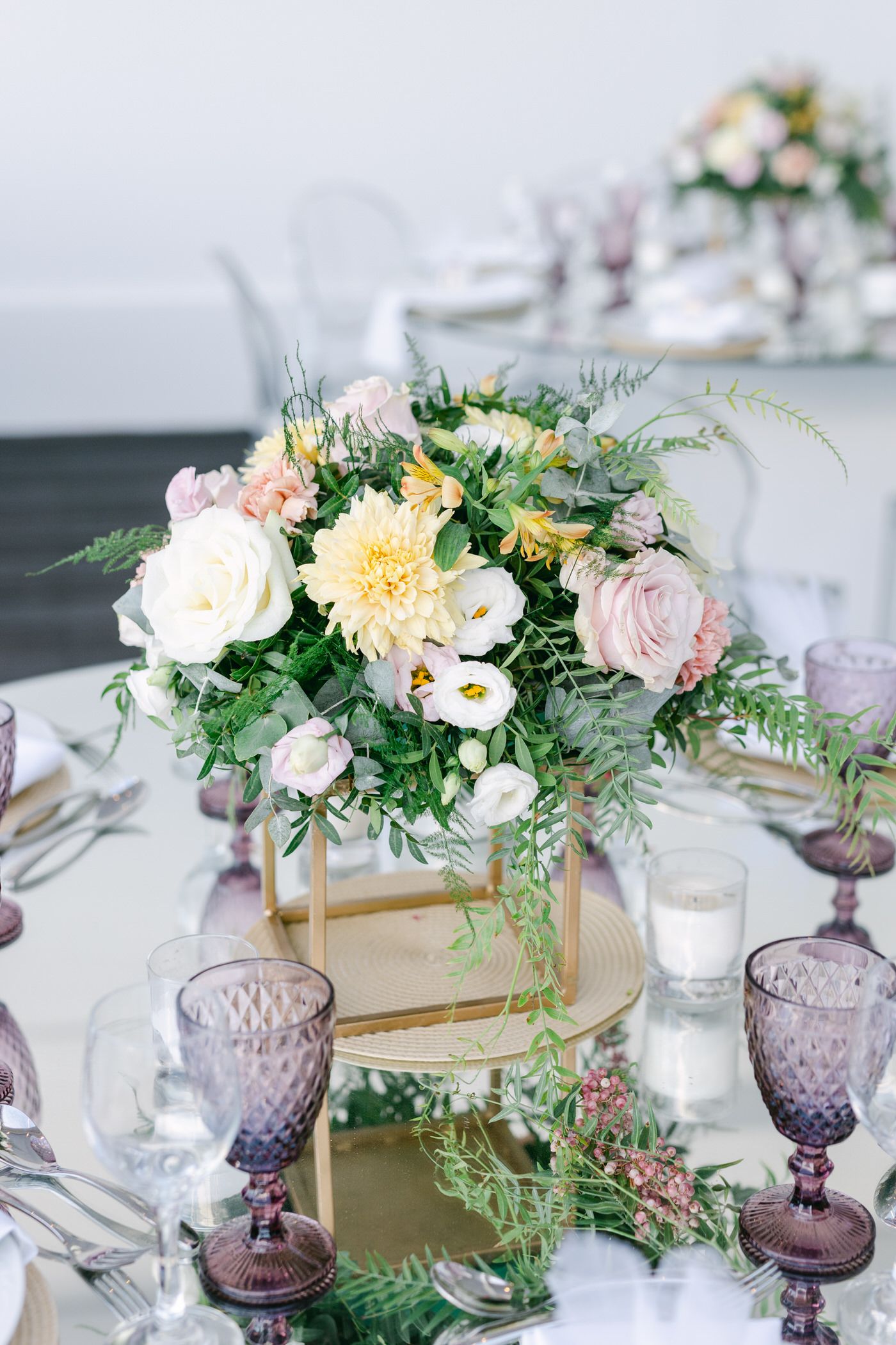 An elegant table setting with purple glassware and a centerpiece of pink and yellow flowers in a gold box vase.