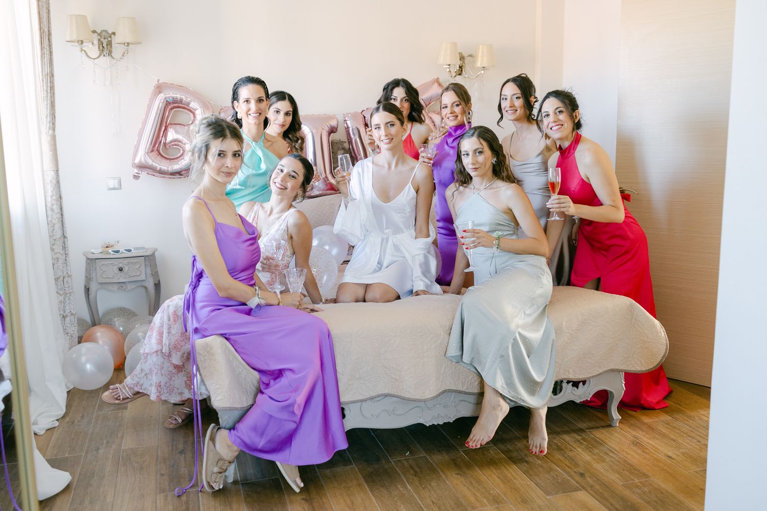 A group in colorful formal dresses sits together on a couch at a celebration with pink balloon decorations.