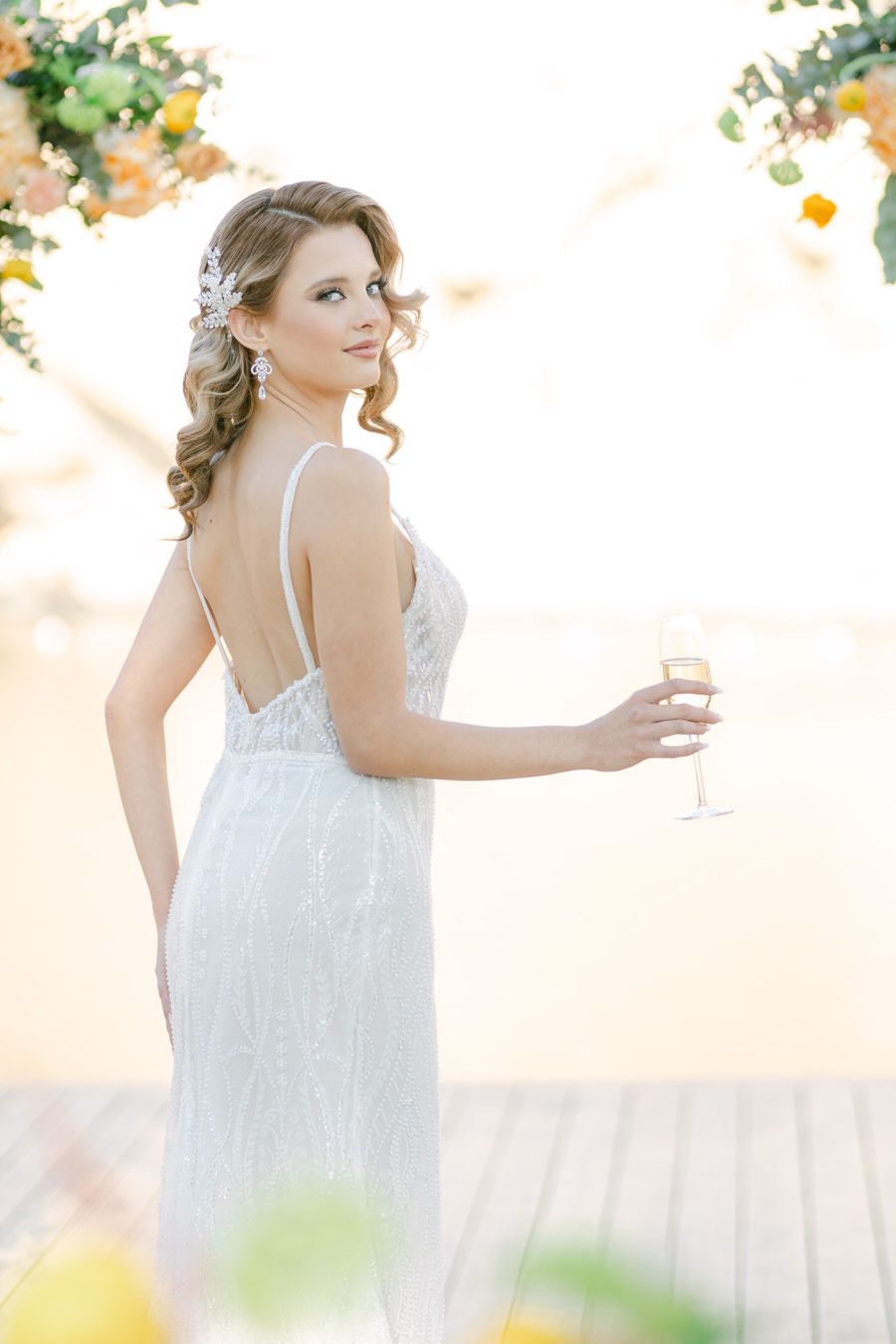 Bride in spaghetti strap gown poses with champagne glass against floral backdrop.