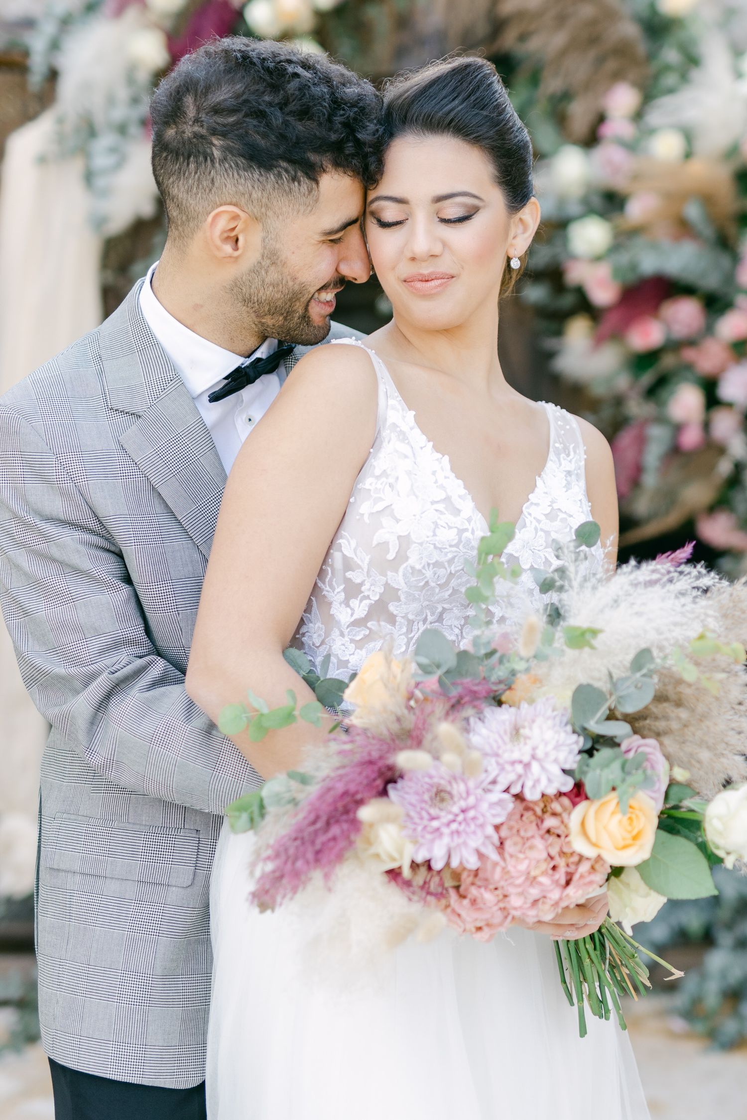 Romantic wedding couple embraces while holding a bouquet of pink and peach flowers.