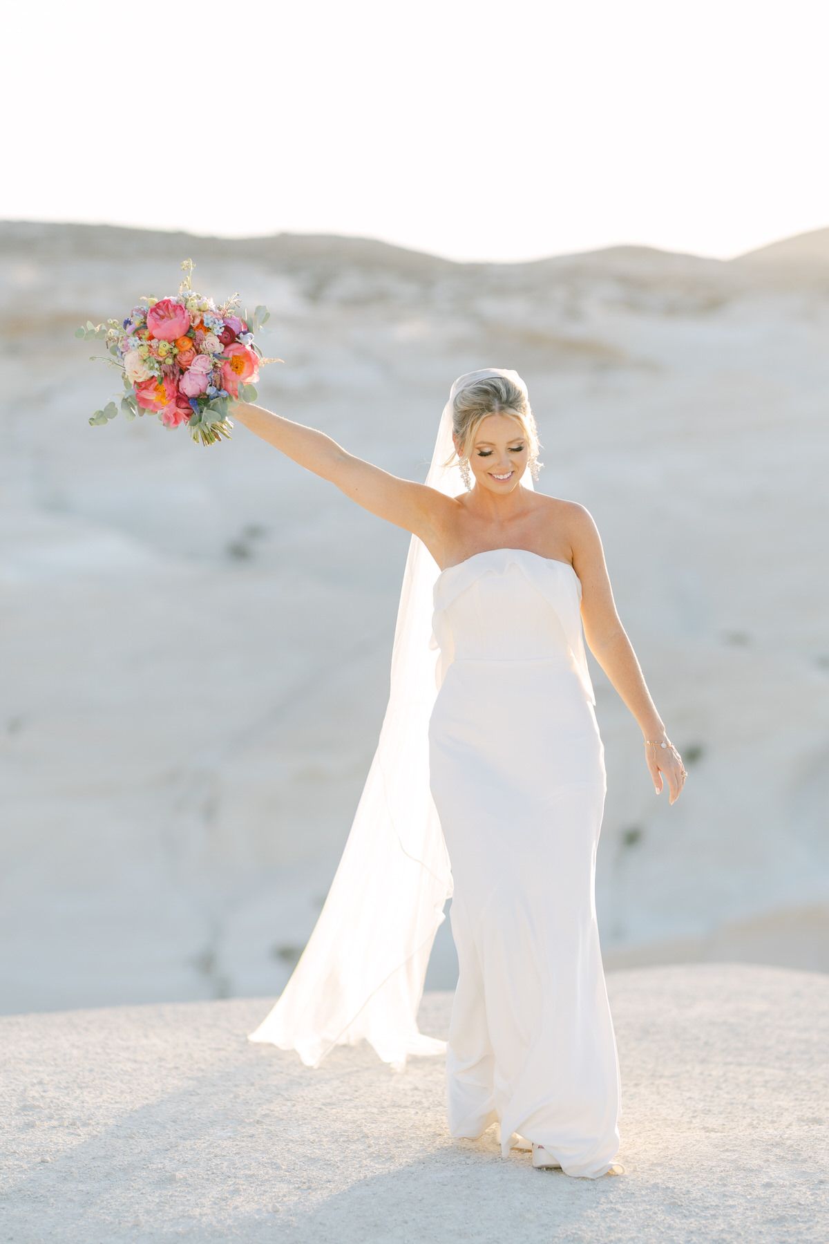 A bride in a strapless white gown holds a vibrant bouquet aloft while standing in sandy white dunes at sunset.