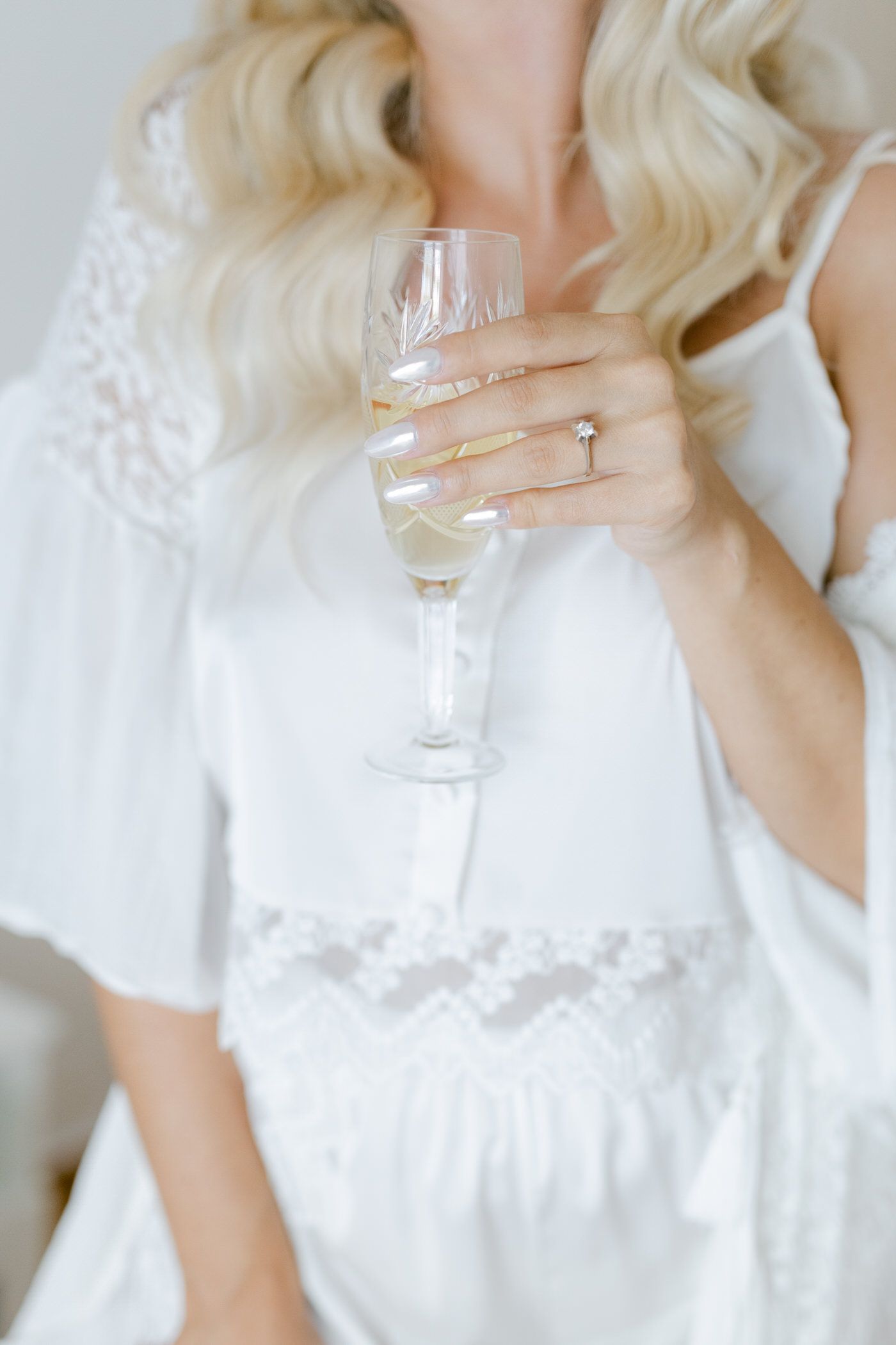 An elegant hand holding a champagne flute shows off a delicate ring while wearing a white lace robe.