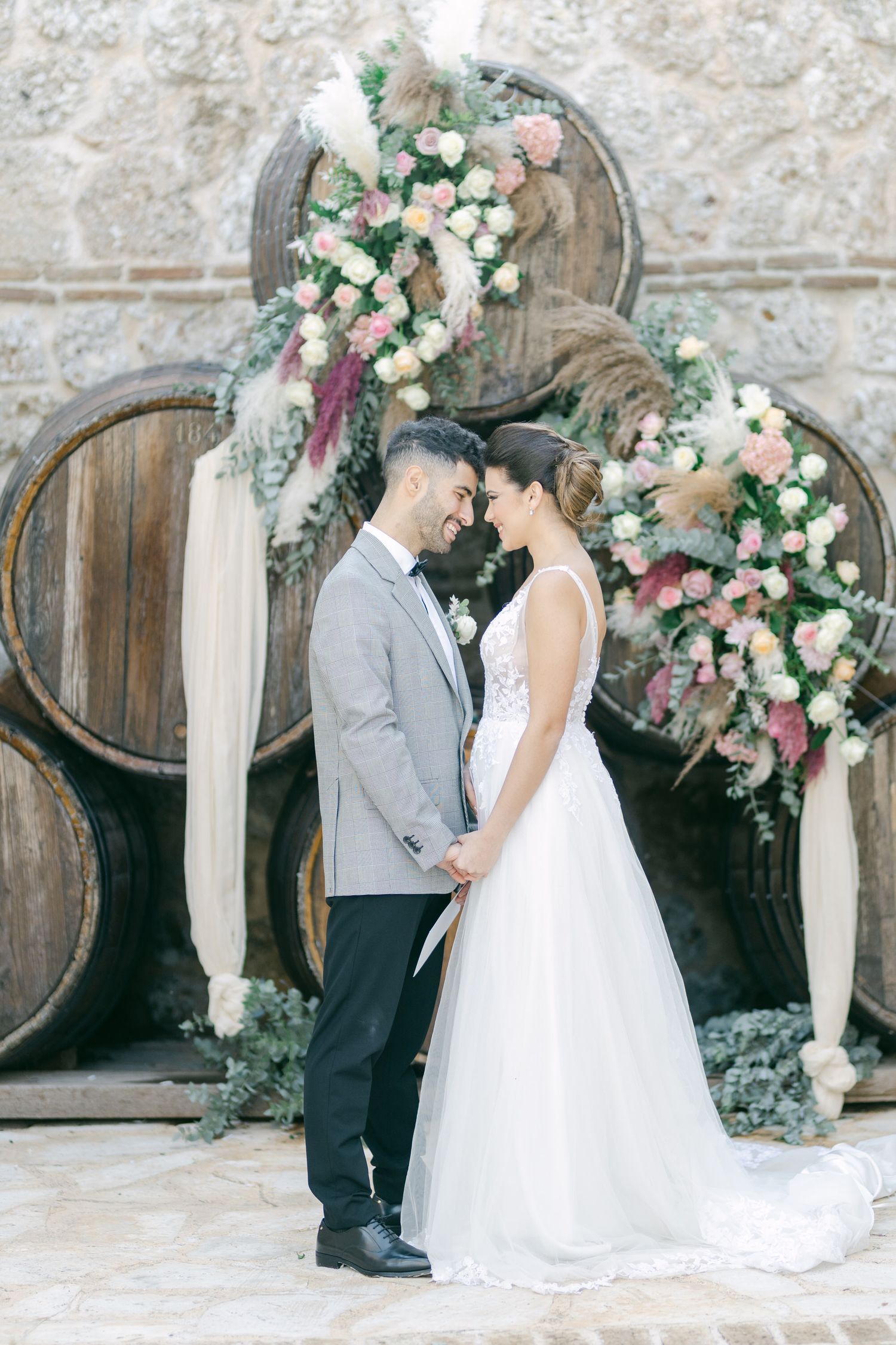 Wedding couple poses in front of vintage wine barrels adorned with pink and white floral arrangements.