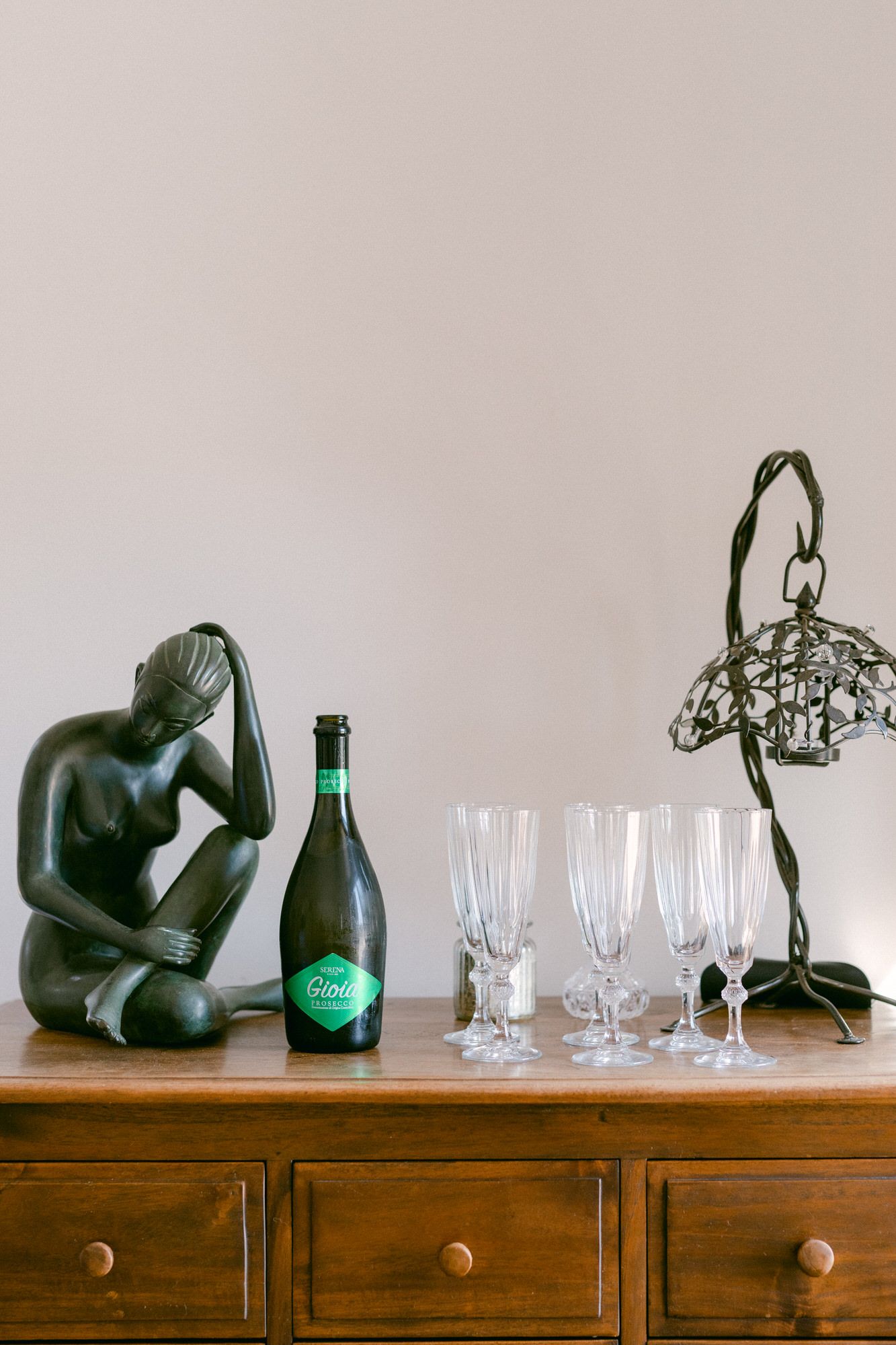 Decorative display with art sculpture, champagne bottle and glasses on wooden dresser.