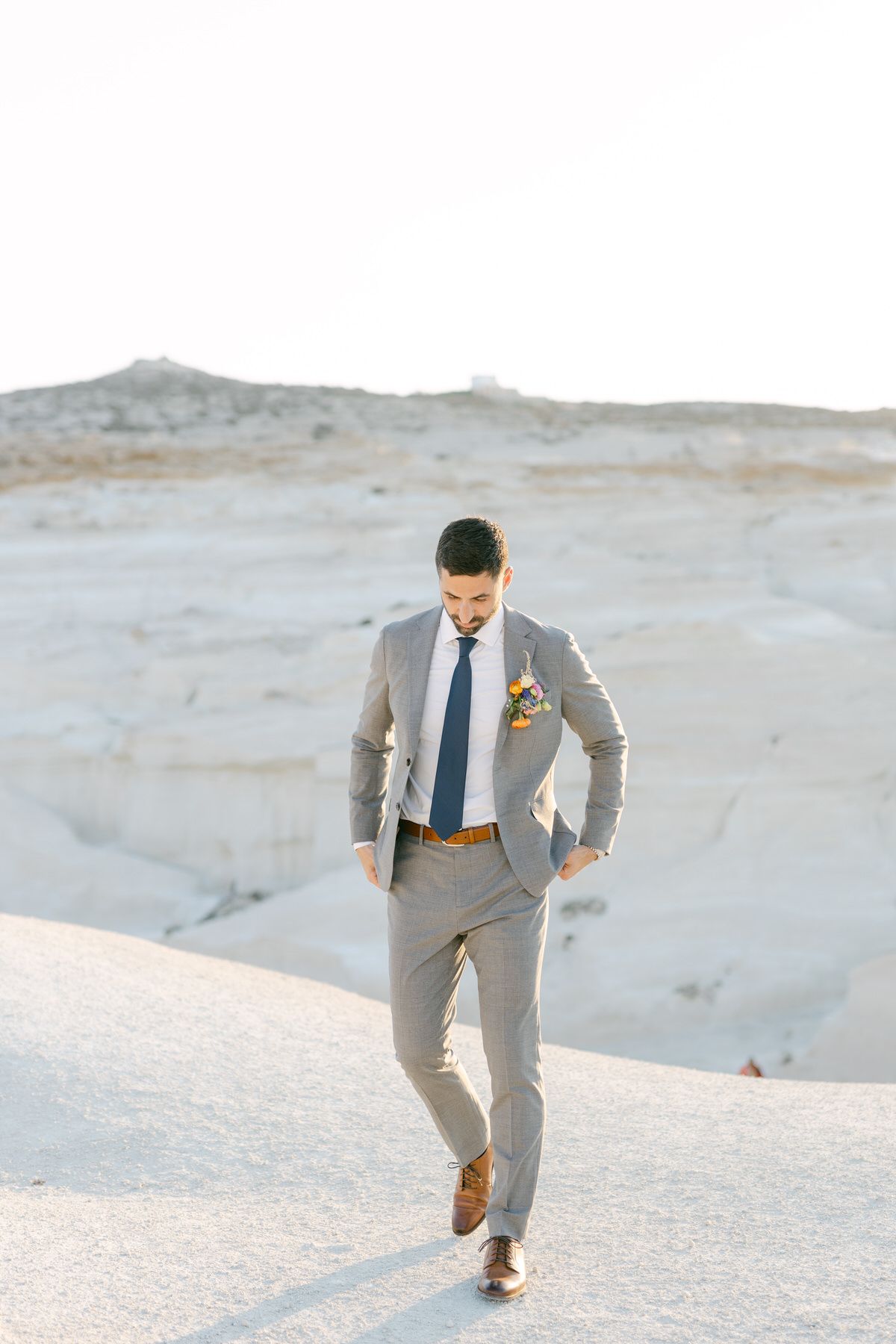 A groom in a light gray suit and blue tie walking down a scenic white desert landscape at sunset.
