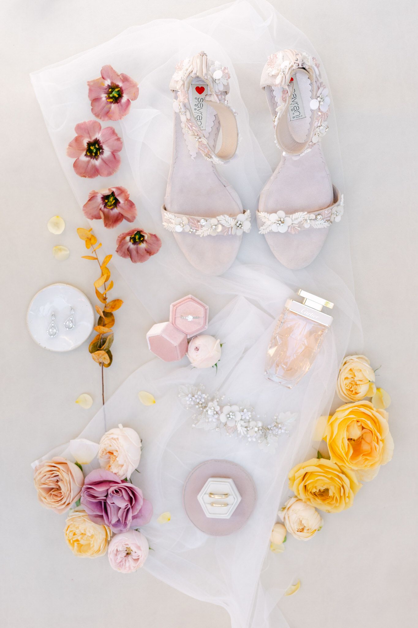 Elegant bridal accessories including pearl sandals, pink and yellow roses, perfume and hair accessories on white background.