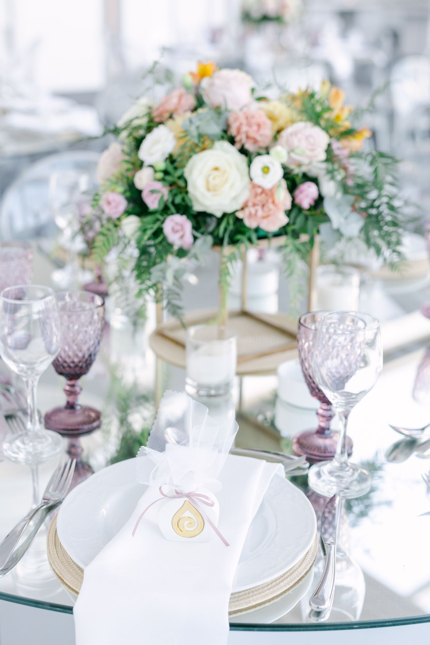 Elegant table setting with purple wine glasses, white plates and blush floral centerpiece in gold vase at wedding reception.