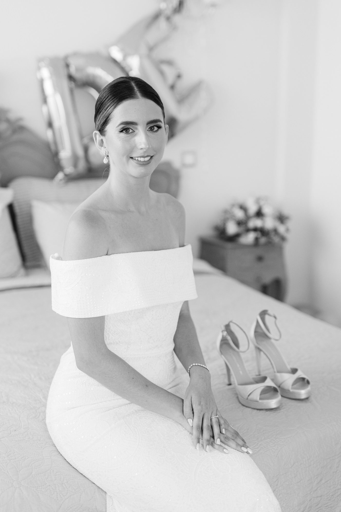 Black and white portrait of a bride in an elegant off-shoulder wedding dress sitting with heels beside her on a bed.