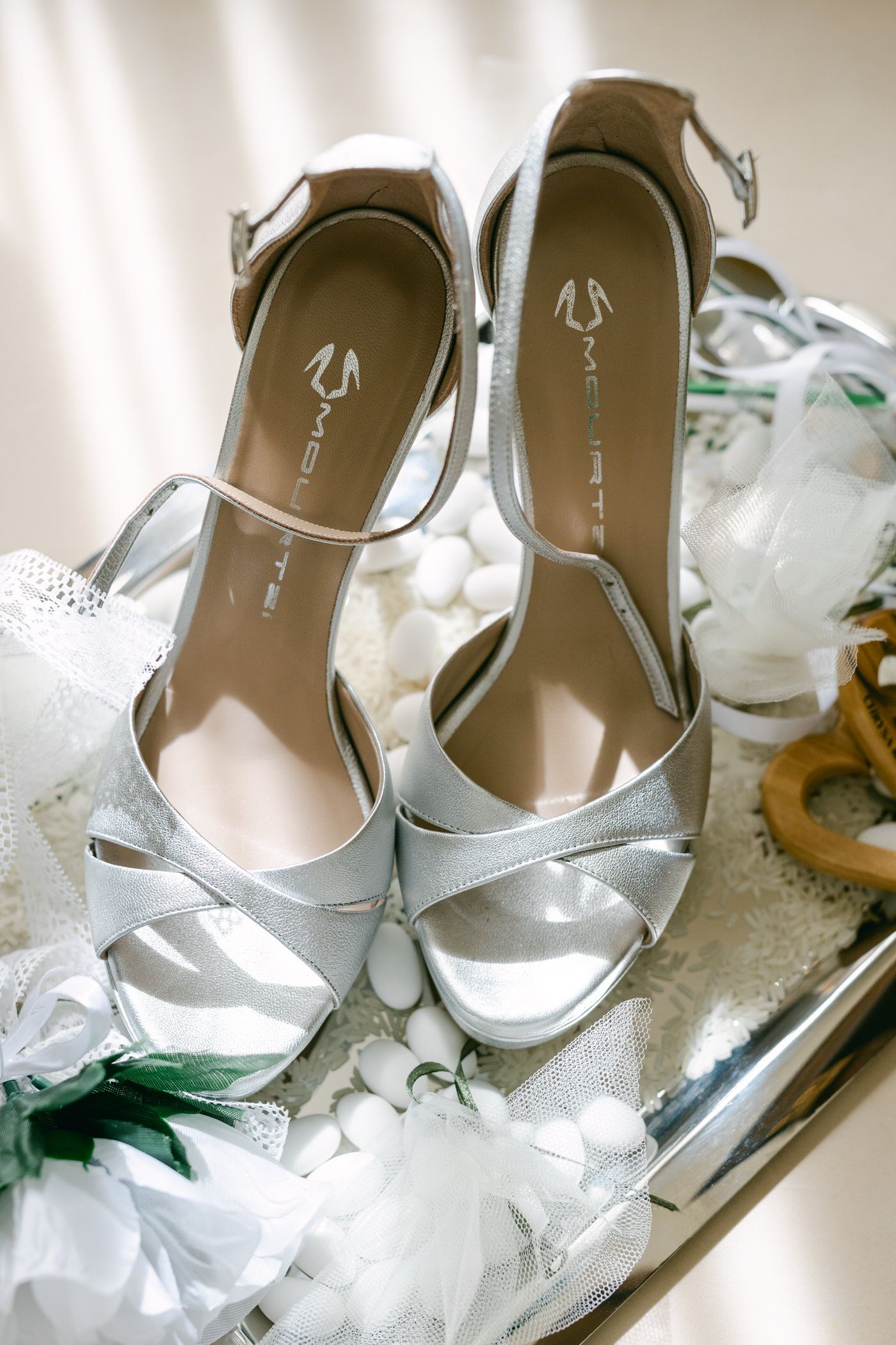 Elegant silver wedding sandals with delicate straps displayed on mirrored surface among white floral decorations.