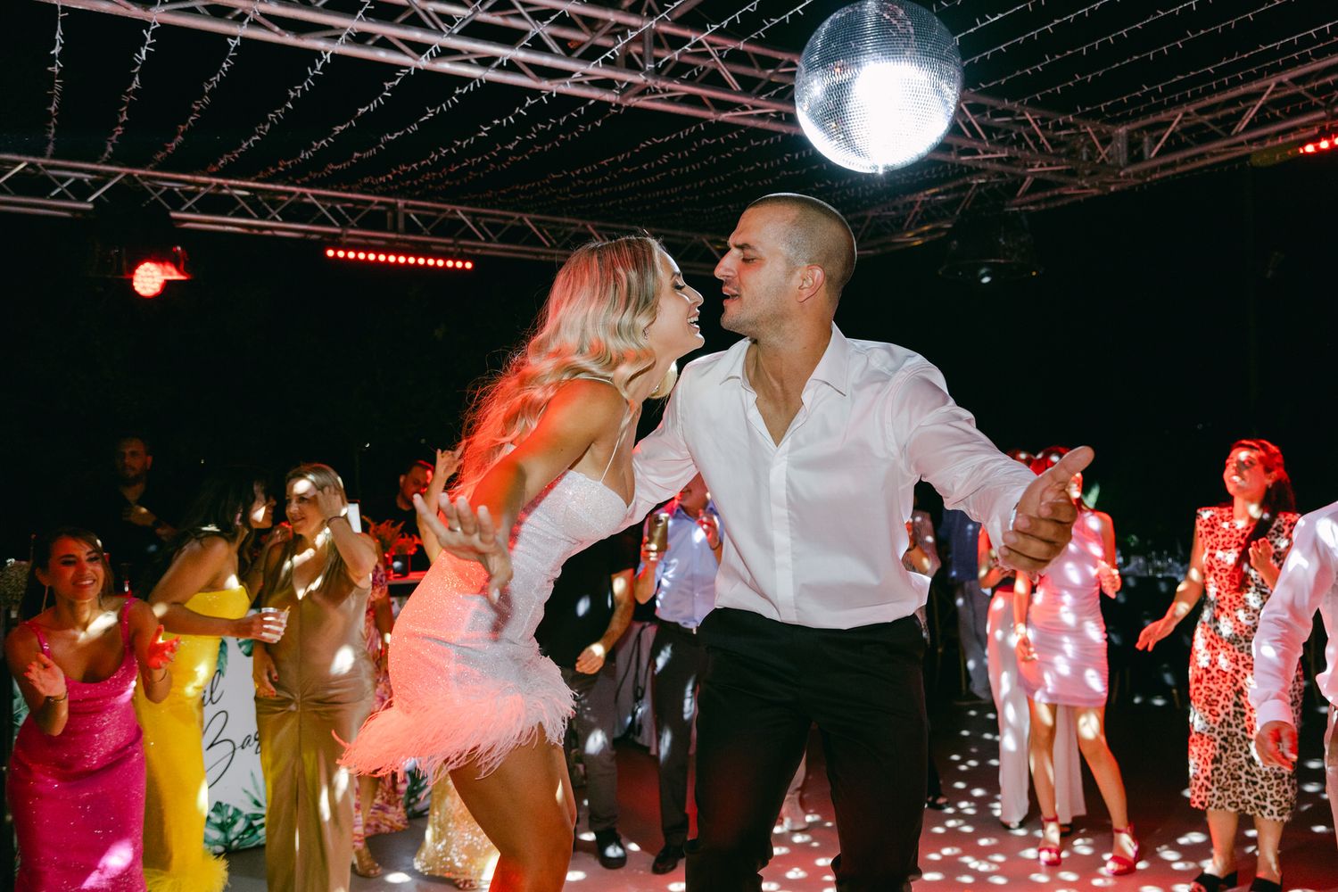 A couple dances intimately under a disco ball on a dance floor surrounded by other guests in colorful attire.