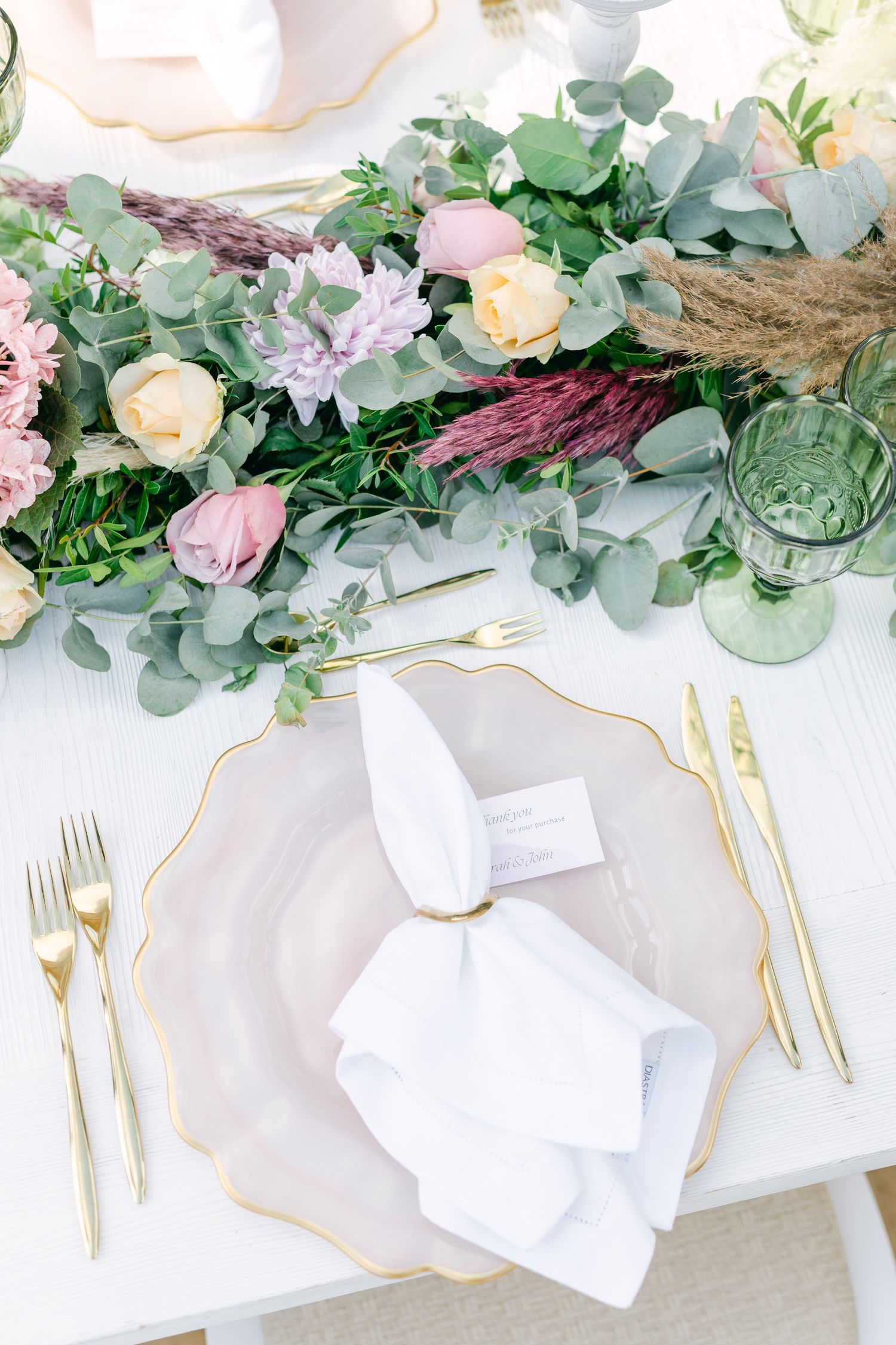 Wedding table decor featuring eucalyptus garland, pink roses, and gold-rimmed place settings.