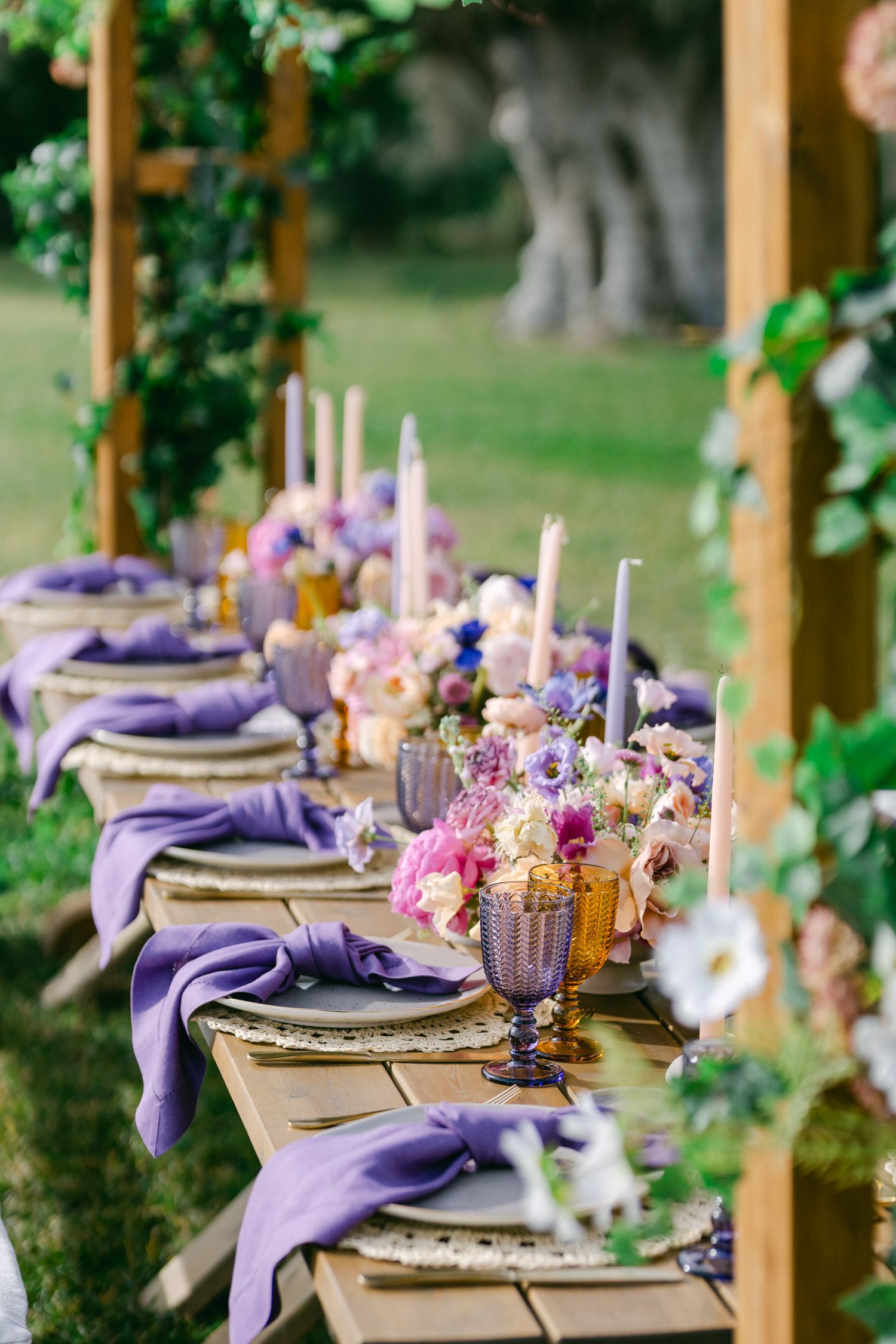 Elegant outdoor table setting with purple napkins and pastel floral centerpieces for a garden wedding reception.