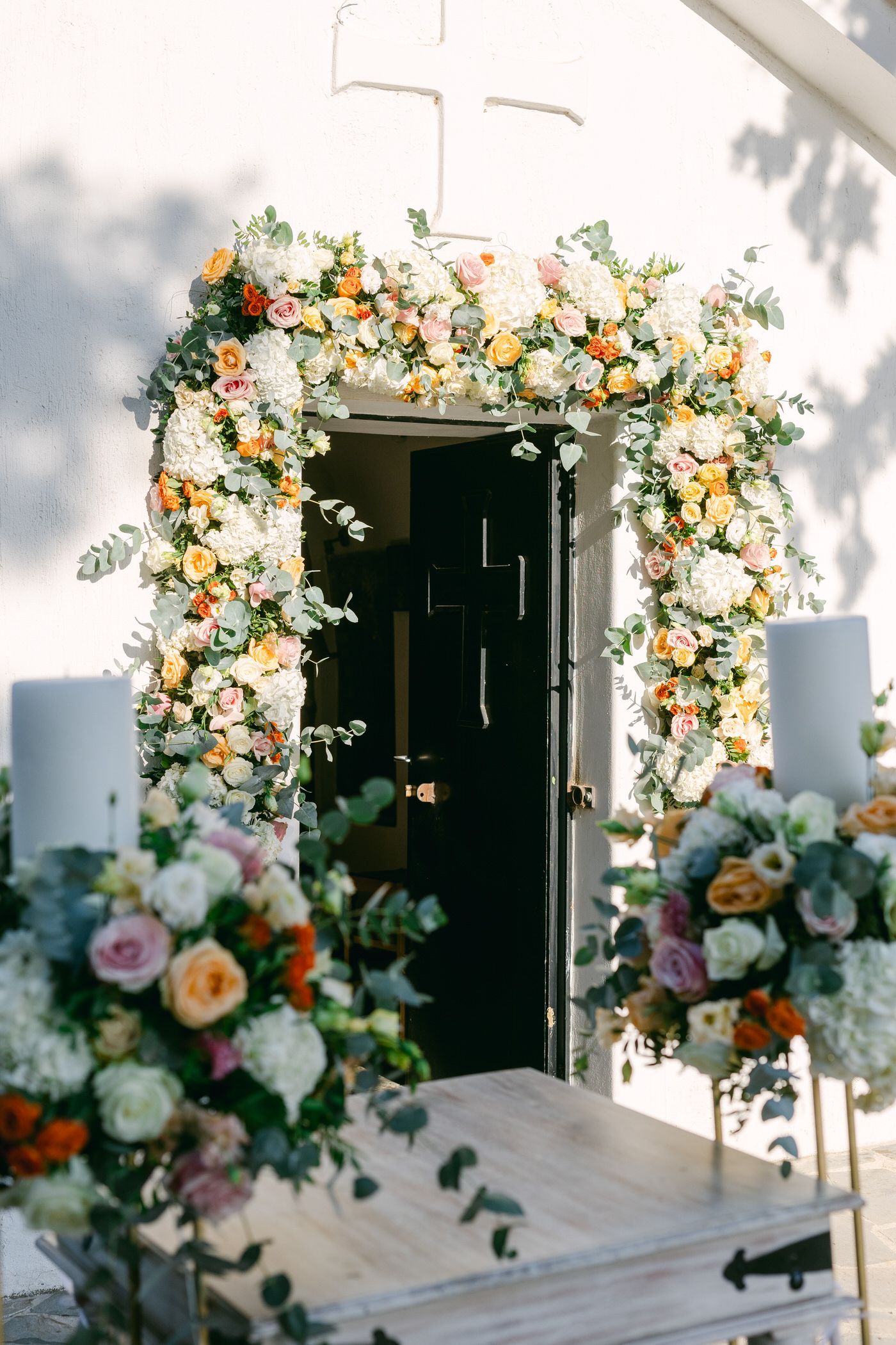 Beautiful floral arch with peach and white roses adorns a church entrance with cross above black wooden doors.