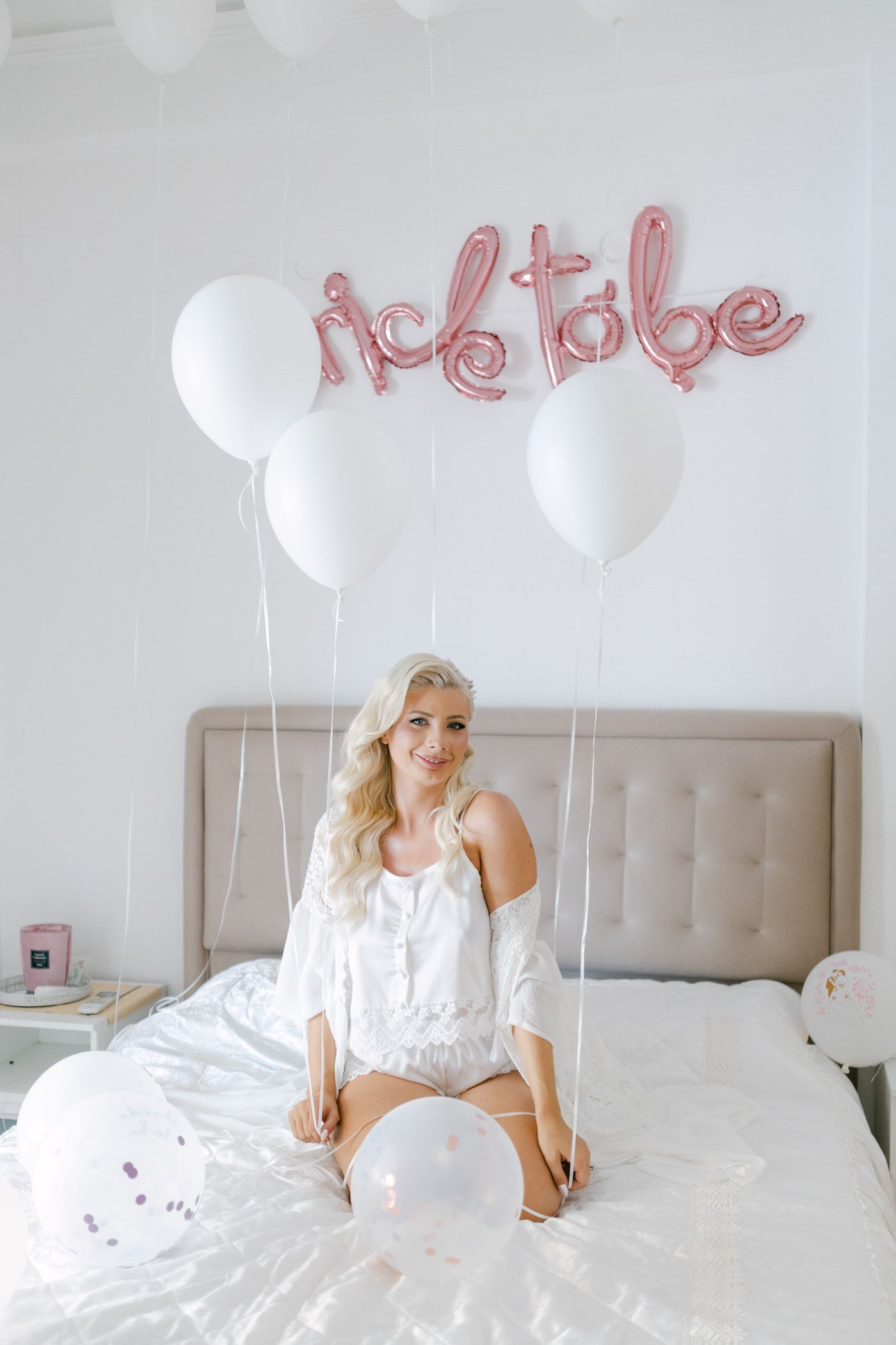 White balloons and rose gold 'bride to be' text decoration float above a bed with white bedding.