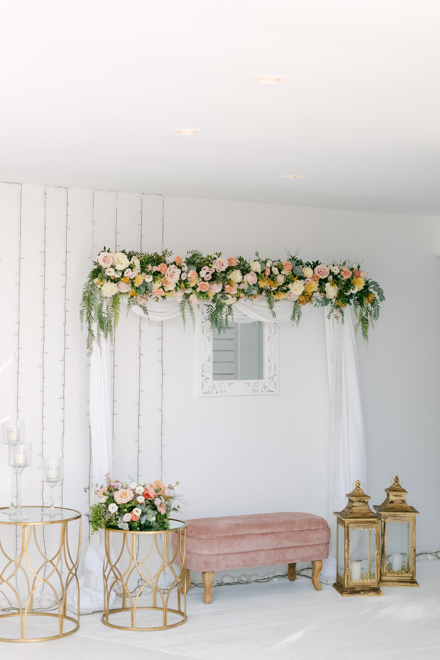Elegant wedding photo backdrop with floral arch, pink velvet bench, gold accent tables and vintage lanterns against white wall.
