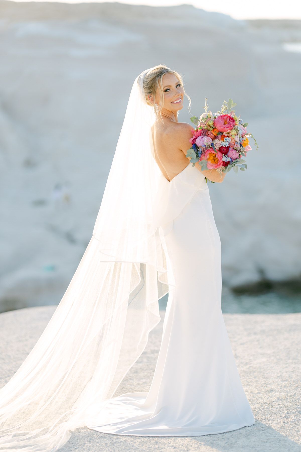 A bride in an elegant white wedding gown with flowing veil holds a vibrant pink and purple bouquet against a light backdrop.