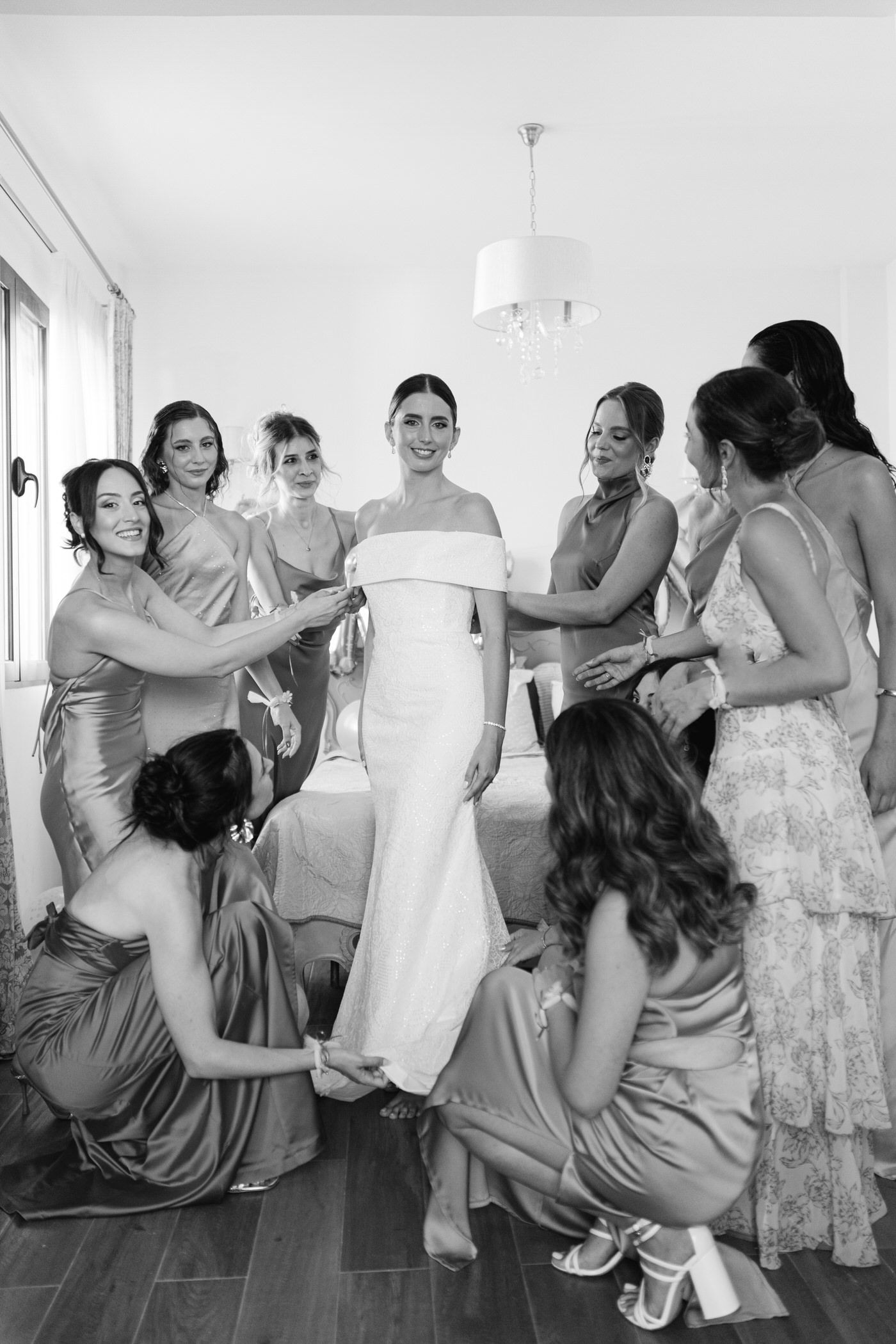 Bridesmaids in pastel dresses help adjust a bride's elegant white off-shoulder gown during wedding preparations.