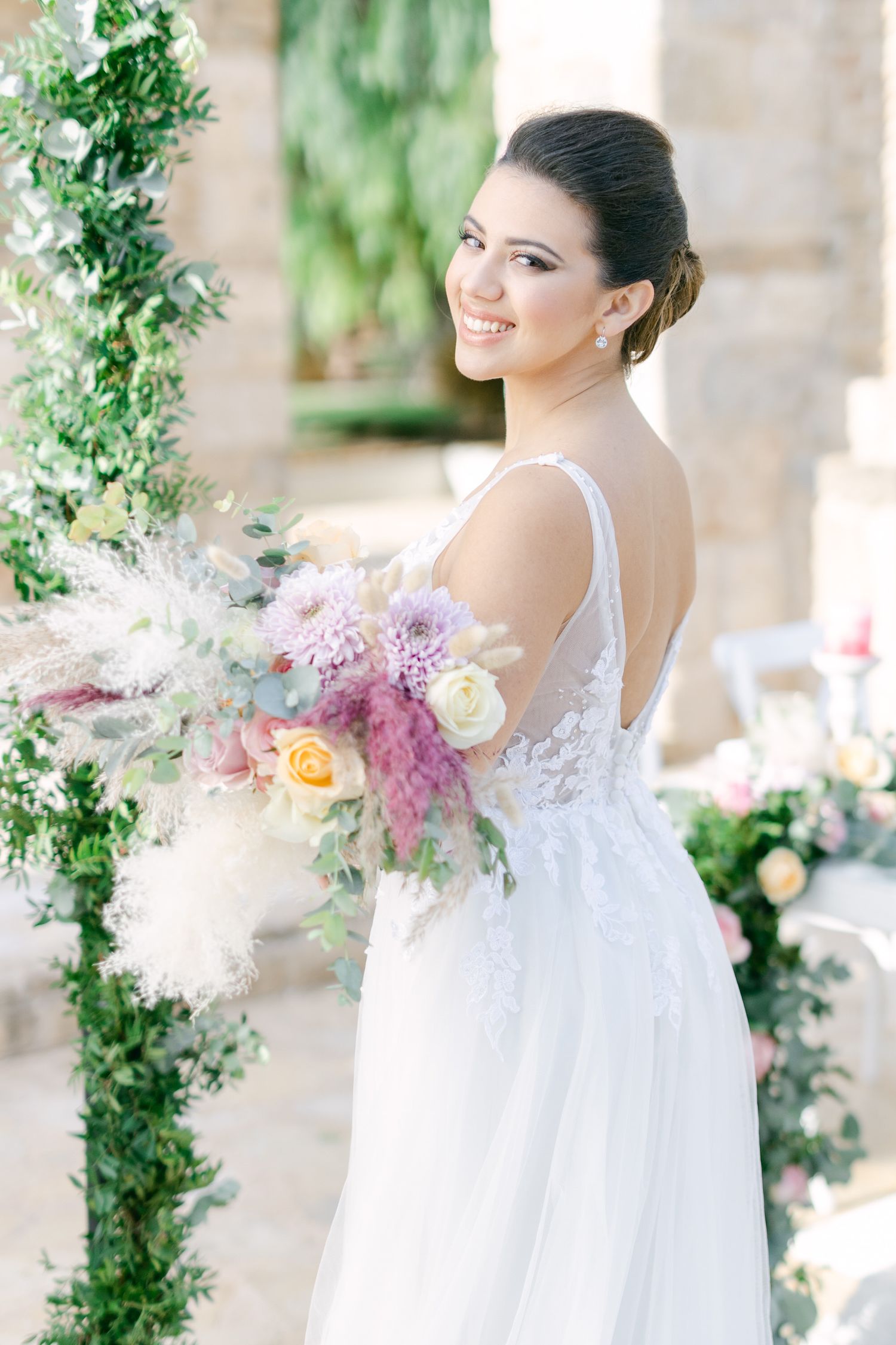 Elegant bride in white lace gown holds pastel bouquet while posing against greenery-adorned backdrop.