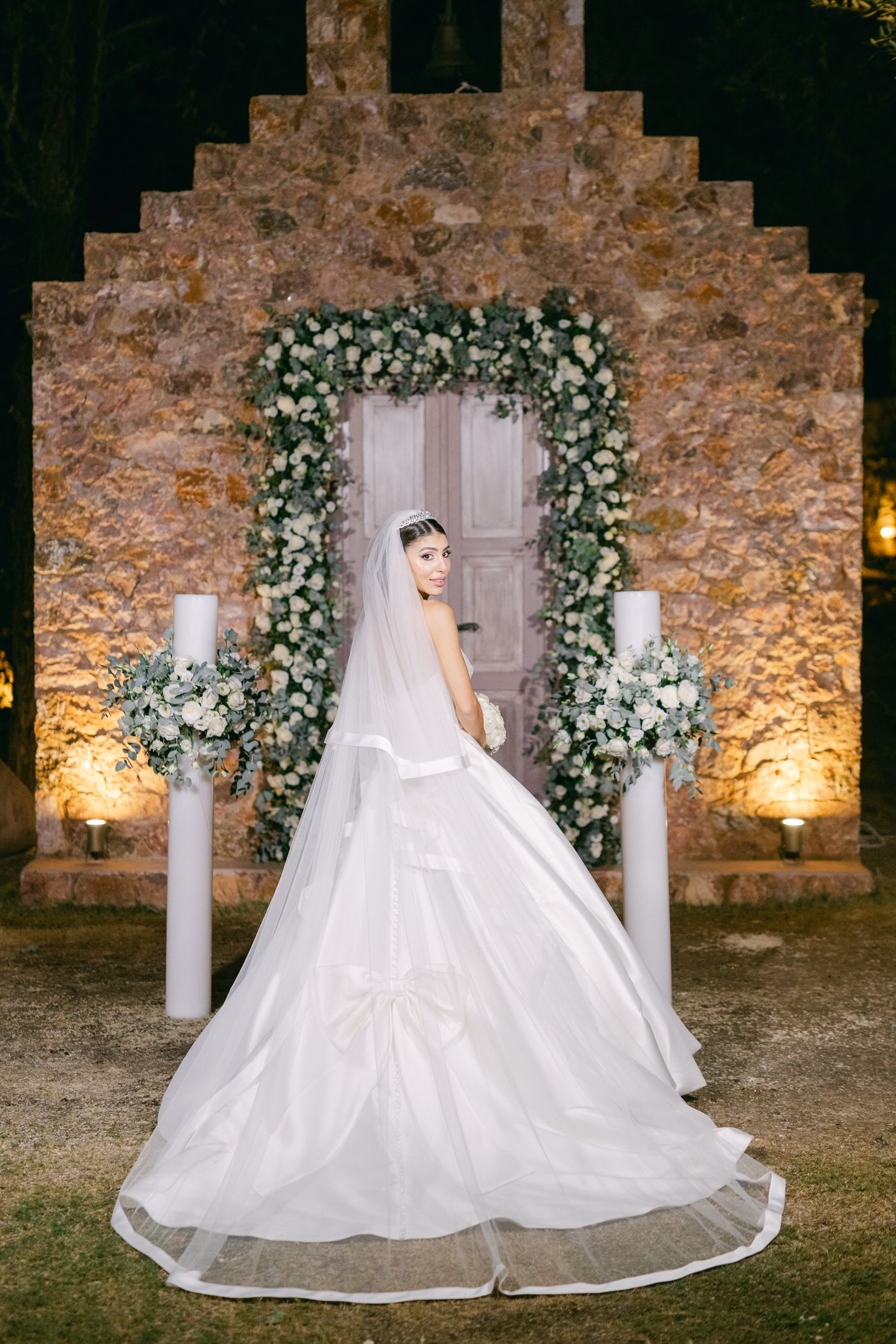 A stunning bridal portrait showcasing a flowing white wedding gown against a stone archway adorned with floral arrangements.