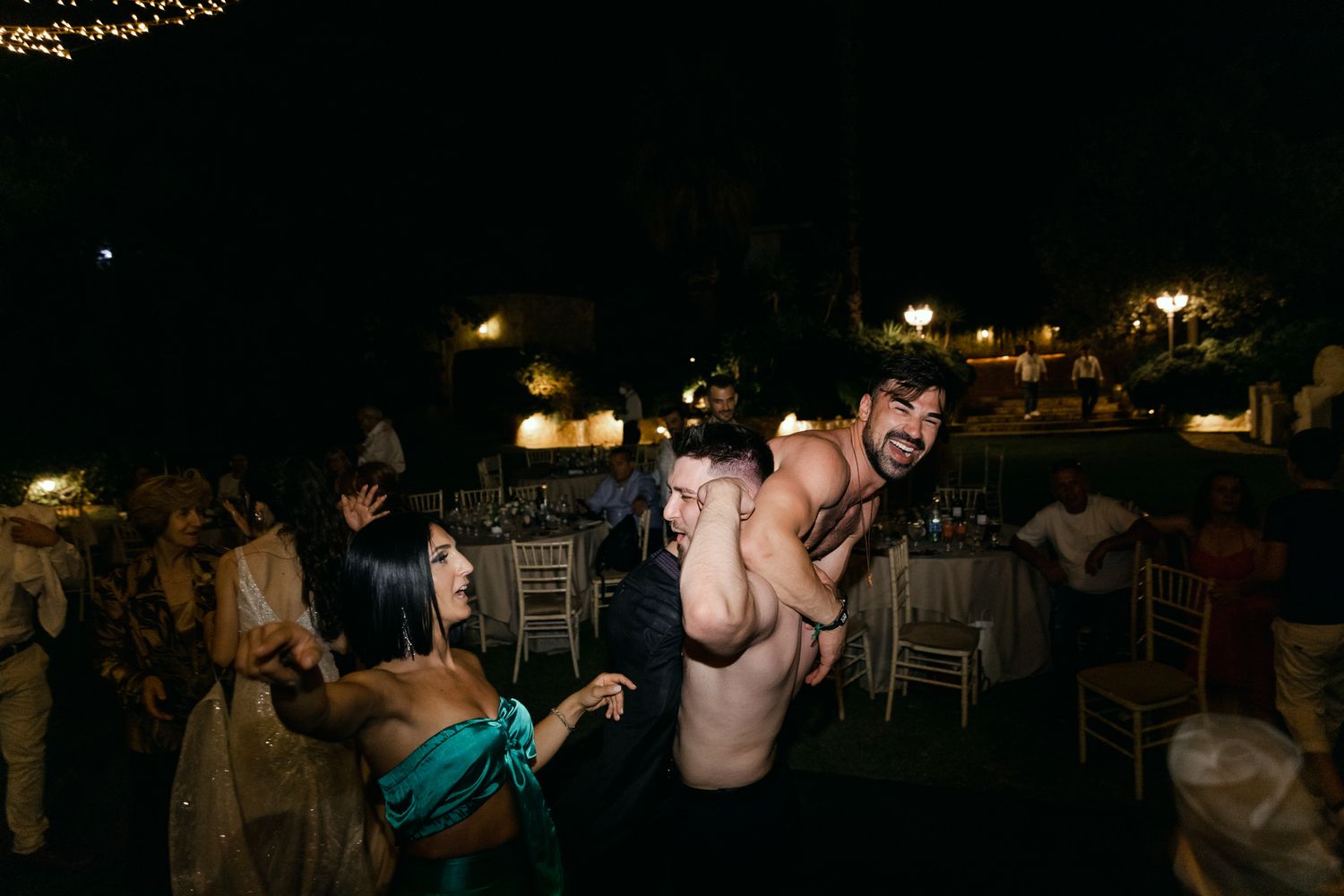 People dancing and celebrating at an outdoor nighttime wedding reception with twinkling lights in the background.