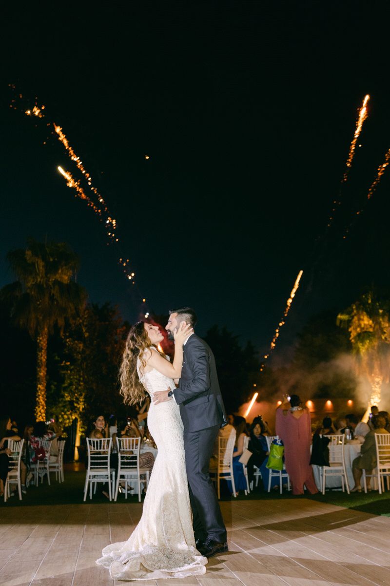 A couple shares a romantic kiss on an outdoor dance floor at night with fireworks lighting up the sky and palm trees in view.