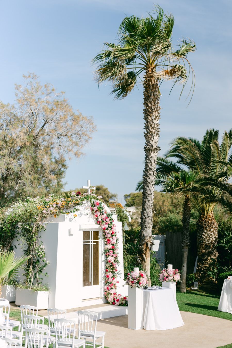 Outdoor wedding venue with white chapel, floral arch, and tall palm trees against a blue sky.