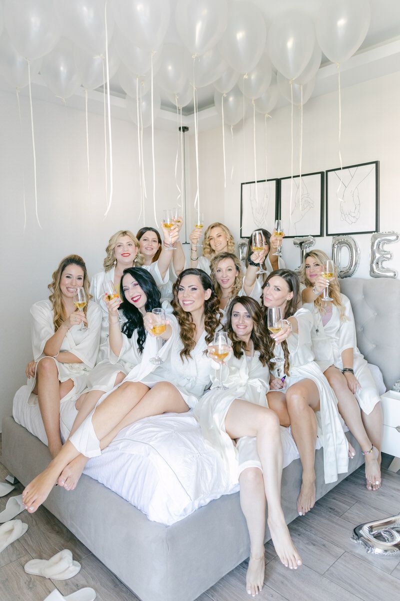 A group in matching white robes sits together on a bed with white balloons floating above during a celebration.