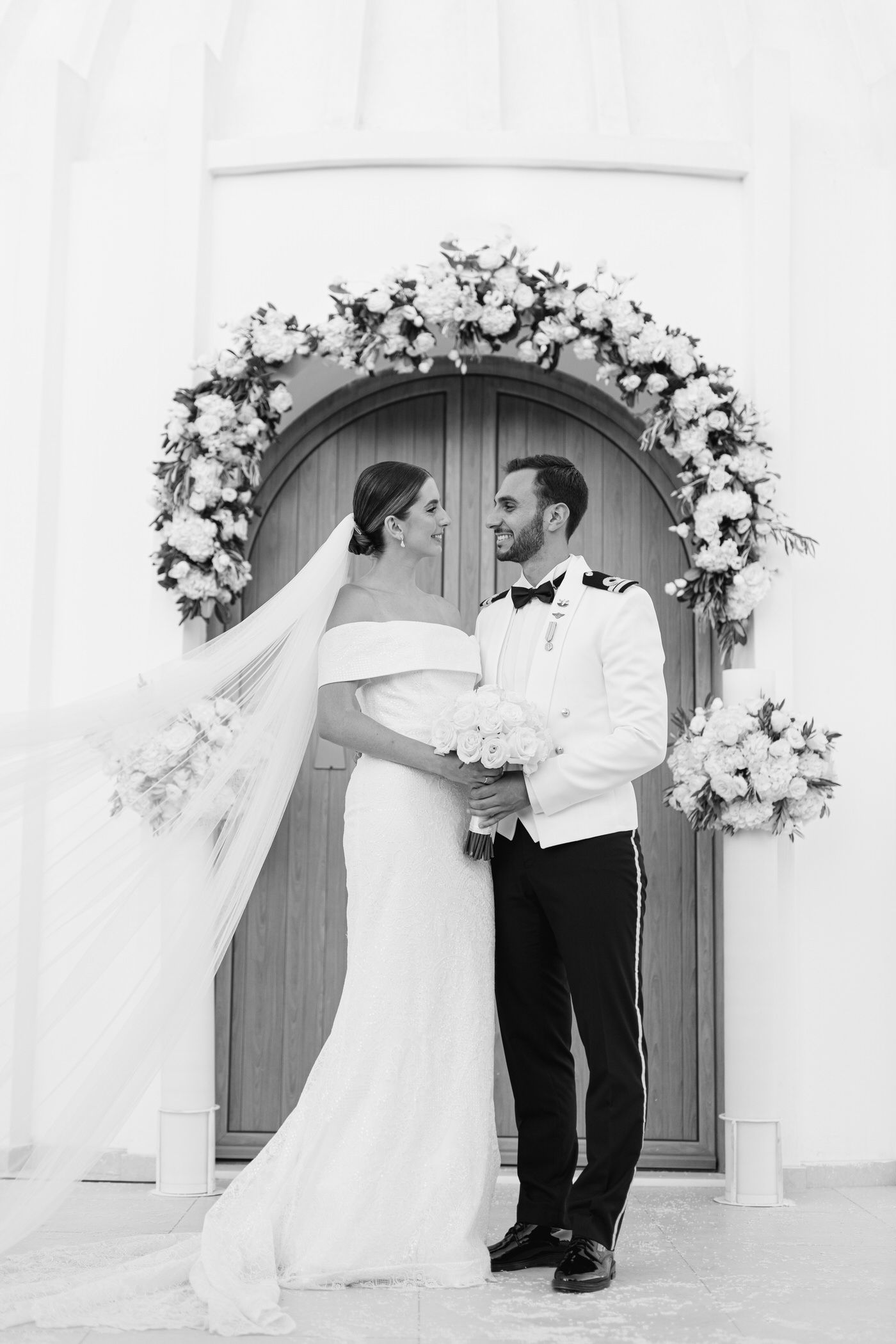 Black and white wedding photo showing couple under elegant floral arch with white flowers and flowing veil.