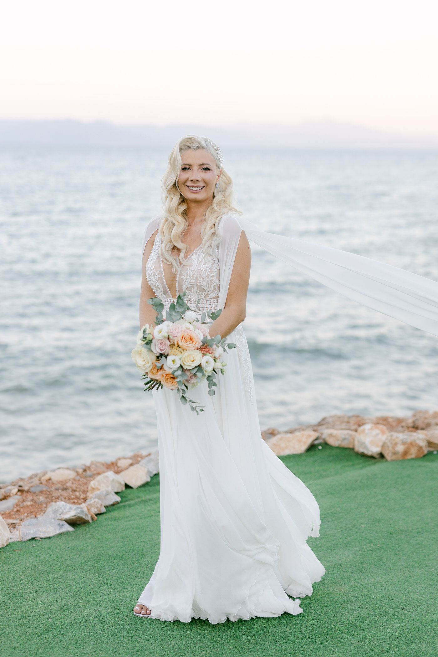 Bride in flowing white lace wedding dress holds peach and green bouquet while standing on grass by ocean cliff.