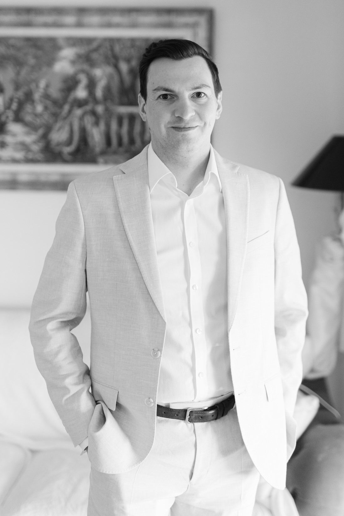 Black and white portrait of someone in a crisp white suit standing casually with hand in pocket against light background.