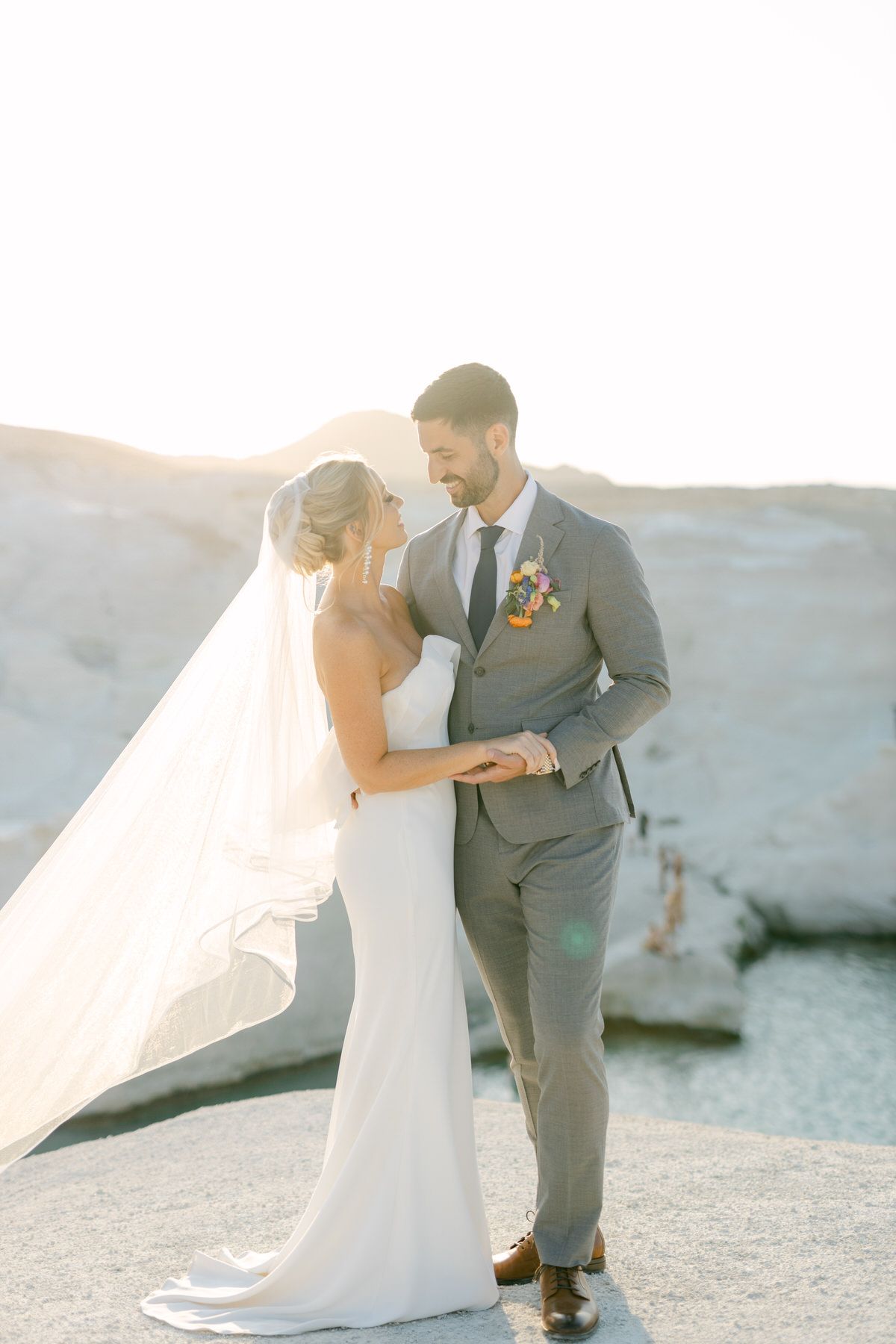 Romantic wedding couple embracing during sunset against white rocky landscape with flowing veil in breeze.