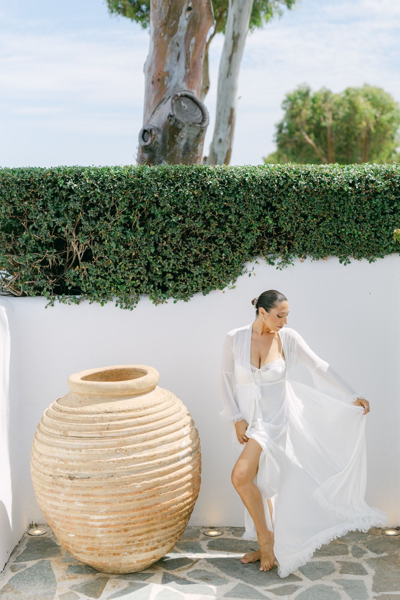 Elegant white summer dress flows in the breeze next to large terracotta pot against Mediterranean-style wall and hedge.
