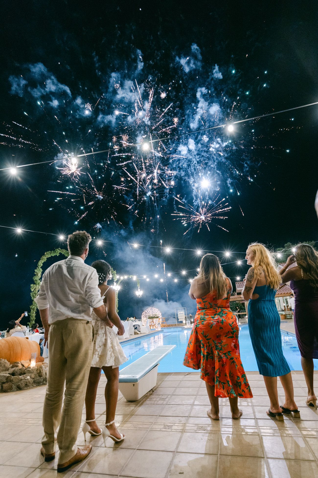 Wedding guests watch blue fireworks display over an illuminated pool at night.